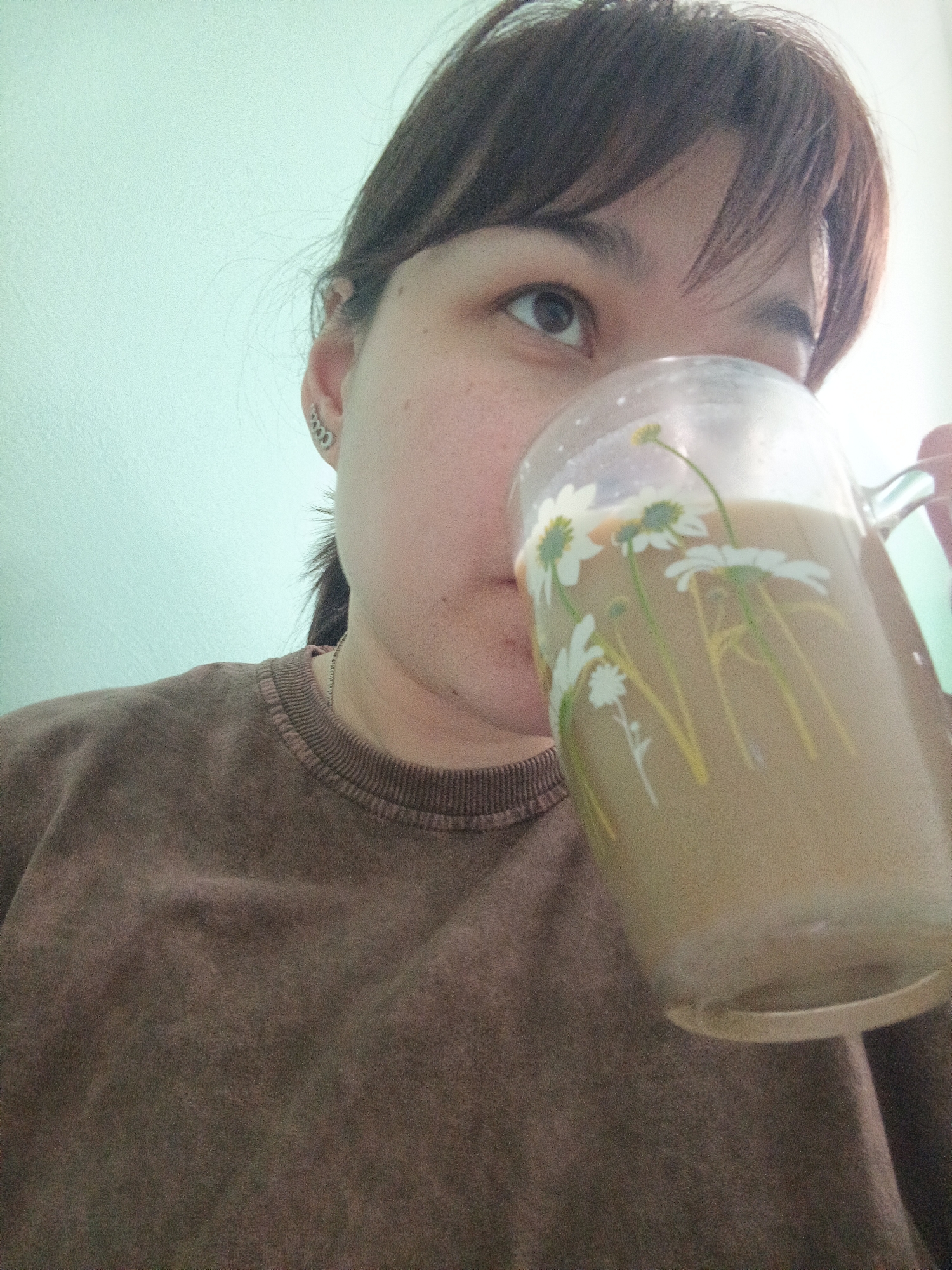 An adult female, with light skin and dark hair, is depicted holding a clear glass mug to her mouth, appearing to drink. She is wearing a dark brown, possibly faded or mottled, crew-neck long-sleeved shirt. A silver-colored ear cuff or multiple linked earrings are visible on her left ear, and a thin silver chain necklace is visible around her neck. Her gaze is directed upwards and to the right. The mug contains an opaque, light brown liquid and is decorated with white and yellow daisy-like flowers with green stems. The background consists of a plain, light blue-green wall. This scene occurs in Chaikovsky, Russia.
