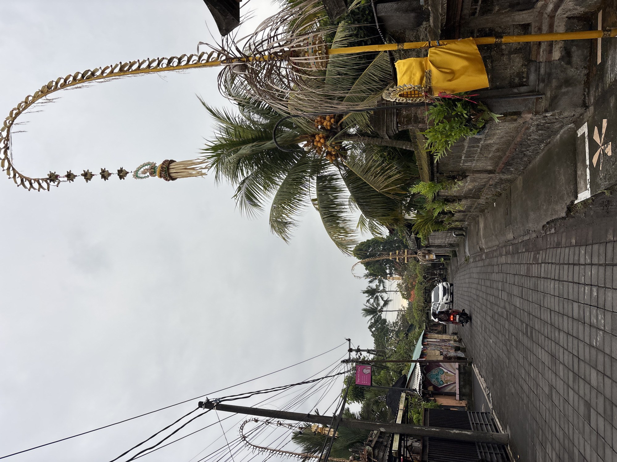 The image depicts a street scene in the Ubud District, Indonesia, on an overcast day. The street is paved with dark gray interlocking bricks and is lined with stone walls and buildings. Overhead, a complex network of electrical wires is visible against the sky. On the right side of the street, a tall, yellow pole is adorned with a large yellow cloth and decorative elements. Further down the street, a white car is parked, and a person is riding a motorcycle, heading away from the camera. Coconut trees with clusters of yellow fruit are scattered throughout the scene, interspersed with other tropical foliage. Elaborate, arched decorations, likely religious or celebratory in nature, are prominent features, one of which extends high into the sky on the right side of the frame, and another smaller one is visible on the left. The buildings lining the street appear to be a mix of traditional and modern styles, with some featuring colorful murals. The overall atmosphere is one of a lived-in, tropical urban environment.