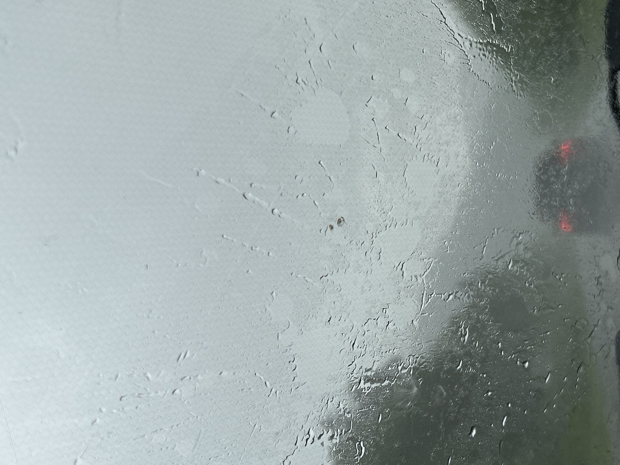 The image shows a view through a wet car windshield, looking out at a hazy, overcast day. The windshield is covered in raindrops and streaks, distorting the view of the outside. In the distance, the taillights of another car are visible, indicating that the vehicles are on a road, possibly in a rural or suburban area. The surrounding environment appears to be green, suggesting trees or vegetation, though the low visibility makes it difficult to discern fine details. The overall impression is one of a dreary, rainy day. There is no indication of the city of Кулмас, Russia, within the image.