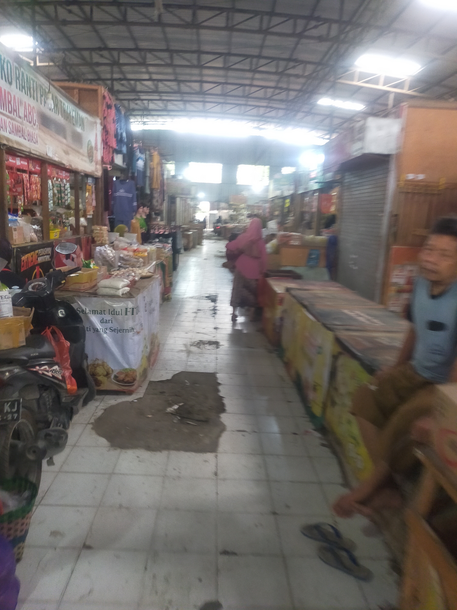 The image depicts a bustling indoor market scene in Trayeman, Indonesia. The perspective is from eye-level, looking down a tiled aisle lined with stalls. The ceiling is a metal structure with translucent panels allowing sunlight to filter through, creating bright areas.
On the left side, a motorcycle is parked, partially obscuring a stall with various goods. A banner on this stall reads "Selamat Idul F... dari ti yang Sejernih" and features images of food. Further down the aisle, a woman in a pink headscarf and traditional clothing walks away from the camera.
The stalls on the right appear to be selling packaged goods and possibly frozen items, with large coolers or display cases lining the aisle. A man in a light blue vest and brown pants is seated in the foreground on the right, looking towards the camera. His body is partially obscured by a stall. There are also scattered items on the floor, including debris and a pair of flip-flops. The overall impression is one of a busy, everyday market environment.
