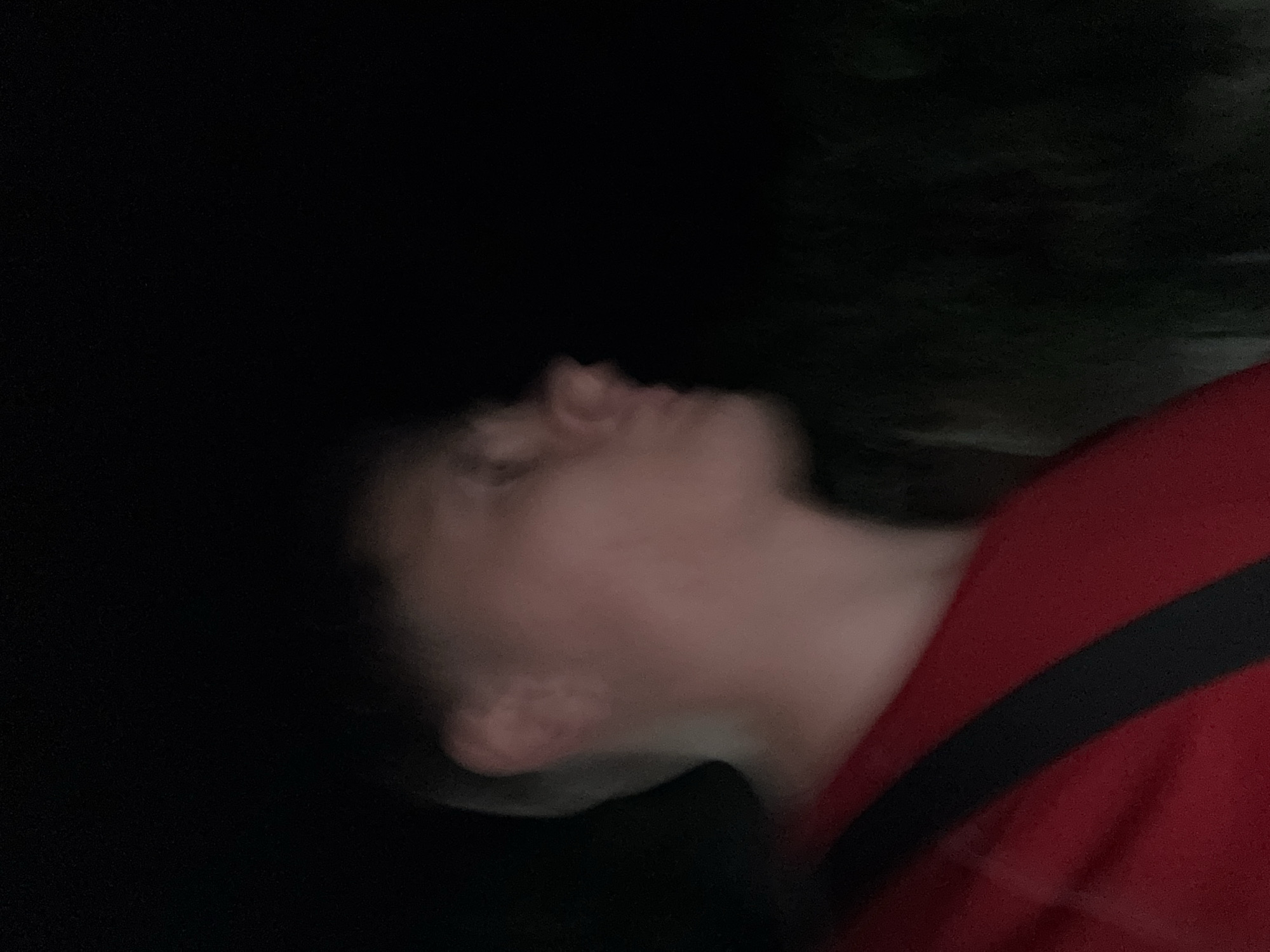 A young person is captured in a low-light, outdoor setting, appearing to be in profile and looking to the right. The person is wearing a red shirt with a dark stripe across the chest. The background is dark and out of focus, suggesting it is nighttime and possibly a natural environment like a park or garden, which could be located in Stara Zagora, Bulgaria. The lighting suggests a flash was used, creating a somewhat stark illumination on the person's face and upper body. The overall atmosphere is intimate and candid due to the shallow depth of field and the subject's profile view.