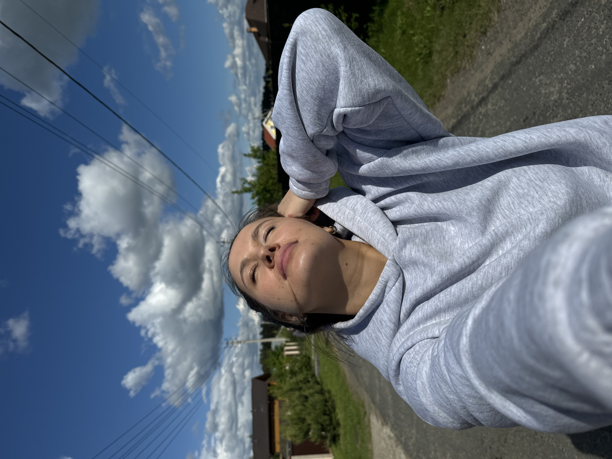A selfie of a young woman with closed eyes, basking in sunlight, is taken outdoors. She wears a light gray hooded sweatshirt. The background shows a blue sky with puffy white clouds and overhead power lines. There is a road and greenery adjacent to a residential area with brown buildings visible in the distance, suggesting a rural setting, identified as Vysokinichi, Russia.