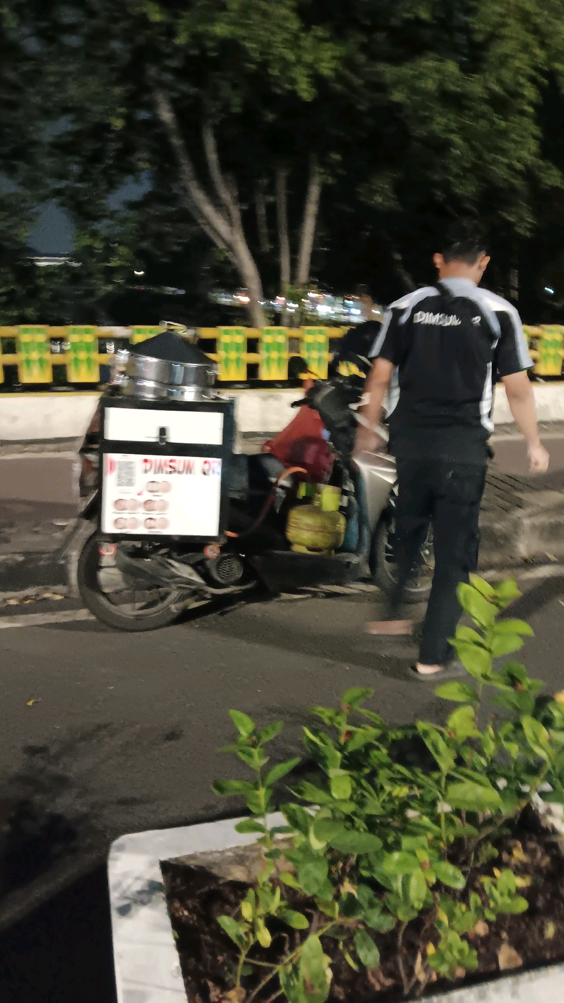 A person wearing a black t-shirt with "DIMSUM Q" printed on the back walks past a parked scooter. The scooter is equipped with a food cart for selling dim sum, featuring a QR code and images of dim sum items. The cart also holds a gas cylinder. The scene appears to be on a street at night, with trees and a yellow and green barricade visible in the background. The location is Central Jakarta, Indonesia.