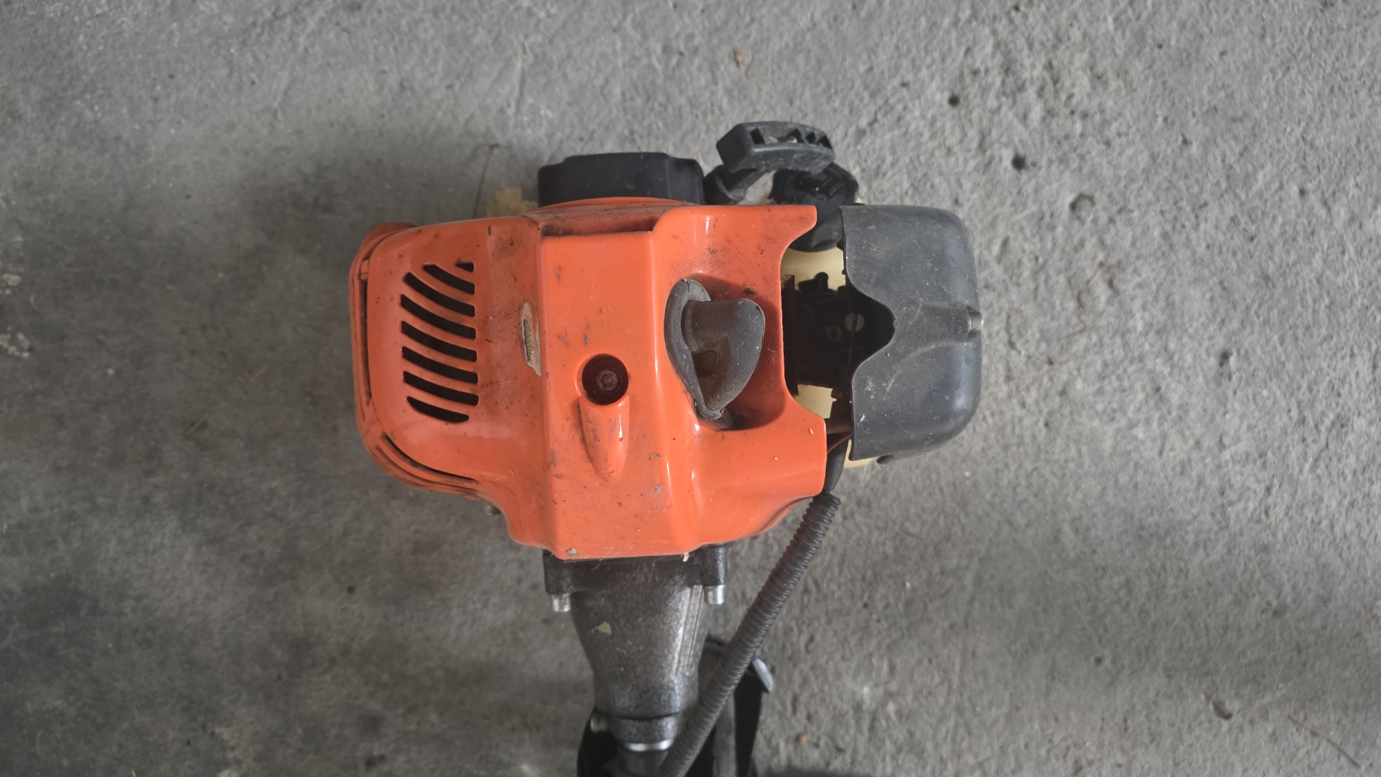 The image shows the engine of a bright orange string trimmer resting on a gray, weathered concrete surface. The engine casing has ventilation slots at the top. A black rubber grip and a black cable are visible, connected to the engine. The background is the unadorned concrete surface. There are no people present in the image. The location is Chuy, Kyrgyzstan.