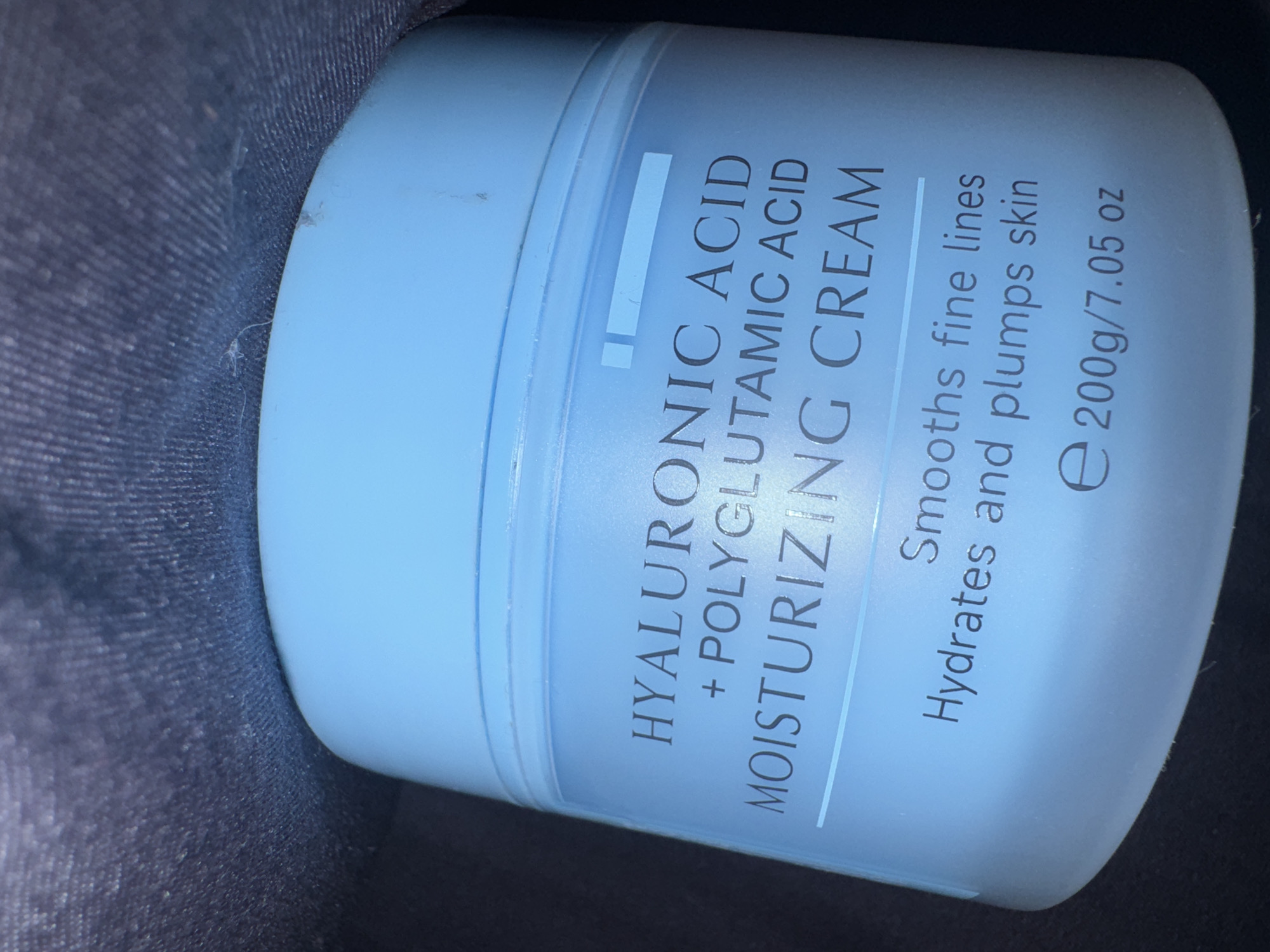 This is a close-up, eye-level, vertical shot of a light blue tub of "HYALURONIC ACID + POLYGLUTAMIC ACID MOISTURIZING CREAM." The tub is placed on a dark, textured surface, possibly fabric like denim, which is out of focus. The lighting appears to be artificial, creating a soft illumination that highlights the texture of the tub and the text.
The text on the tub is clearly legible and describes the product's benefits: "Smooths fine lines Hydrates and plumps skin." It also indicates the product's weight: "e 200g/7.05 oz". The overall impression is one of a skincare product being showcased, with emphasis on its ingredients and intended effects. There are no people or specific environmental cues to suggest a particular time of day, weather, or location beyond the presence of the product itself. The background is dark and undetailed, keeping the focus squarely on the cream tub.