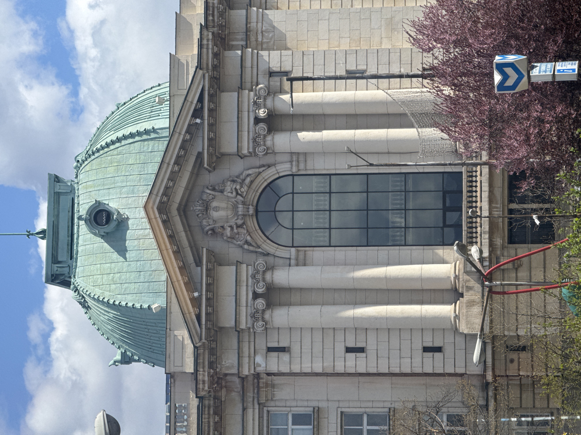 A vertical, medium shot depicts a section of the Central Mineral Baths building in Sofia, Bulgaria. The building features a prominent green copper dome on the left side of the frame, partially obscured by the blue sky with white clouds. The building’s facade is pale stone, decorated with classical architectural elements, including columns with Corinthian capitals, entablatures, and a large arched window. A sculpted face adorns the area above the window. A purple-leafed tree with a metro sign is on the right side of the building.