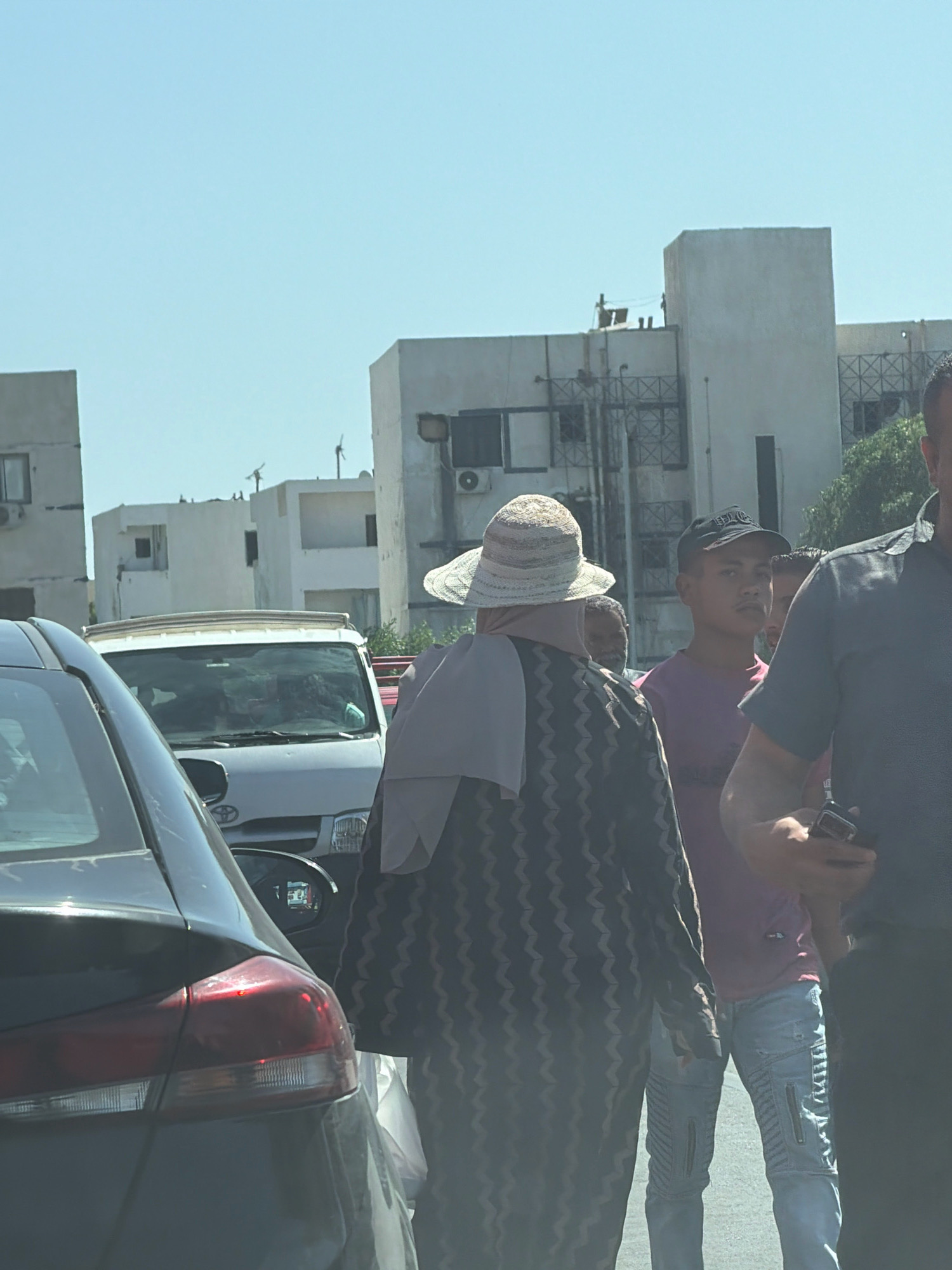 The image captures a street scene in Sharm El Sheikh, Egypt, under a clear blue sky. In the foreground, the rear left of a dark-colored car is visible. Beyond this car, a white van with its roof rack exposed is parked. Several people are present in the middle ground and background. A woman wearing a patterned dress, a light-colored wide-brimmed hat, and a grey hijab is facing away from the camera. To her right, a young man in a purple shirt and a black baseball cap is looking towards the camera. Another individual is partially visible behind him. To the far right, a person wearing a dark grey shirt is holding a mobile phone and looking down. In the background, there are multiple white buildings with varying architectural styles, some featuring window frames and visible air conditioning units. The overall impression is of a typical urban street during daylight hours.