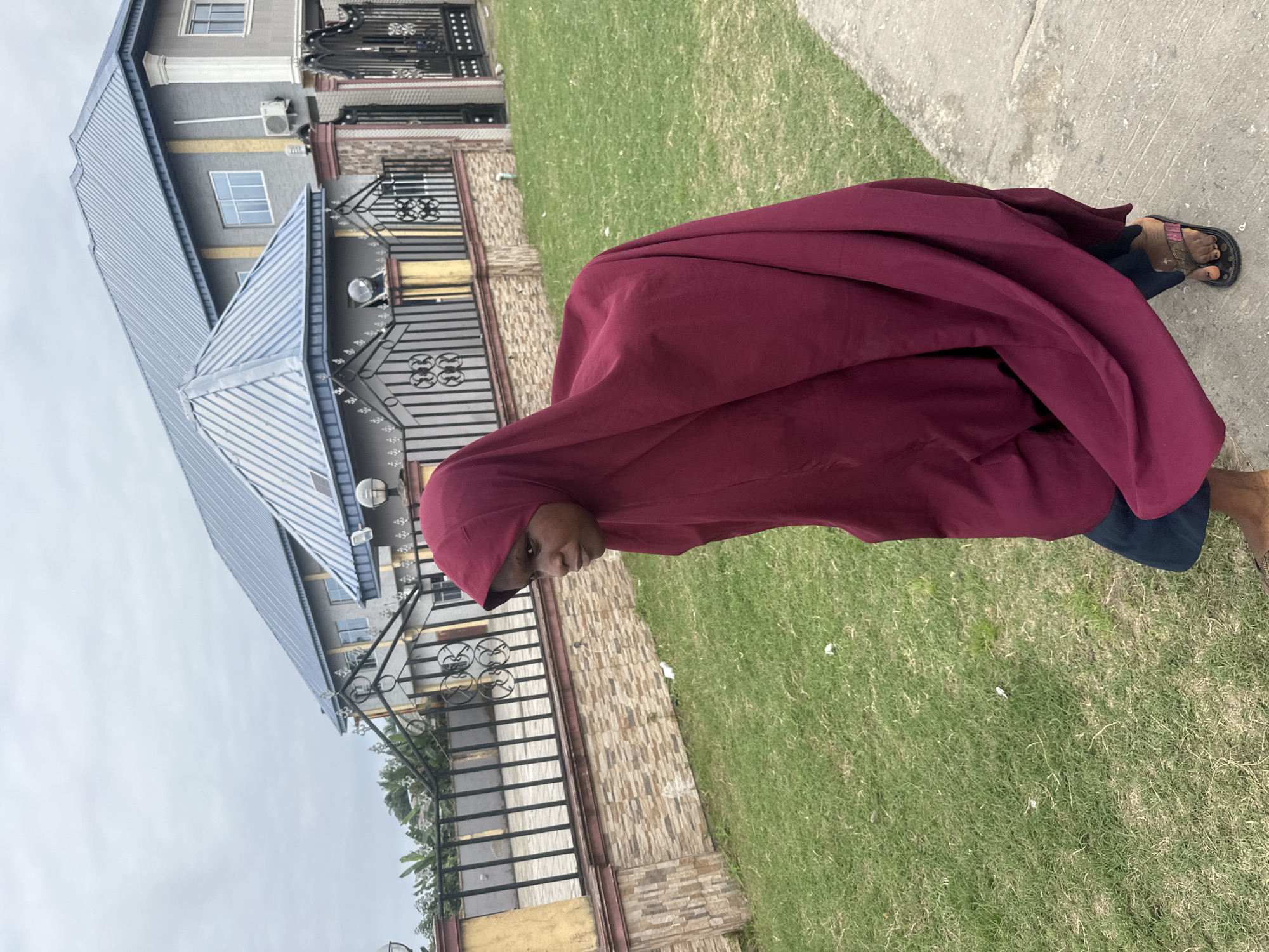 A young female, wearing a maroon hijab and dark trousers, walks on a concrete sidewalk next to a grassy lawn. Behind the lawn is a low stone and metal fence, and beyond that, a grey building with a blue metal roof. The building has multiple windows and an air conditioning unit visible on its exterior. The scene appears to be located in Badagry, Nigeria, given the architectural style and lush vegetation. The woman is looking off to the left side of the frame.