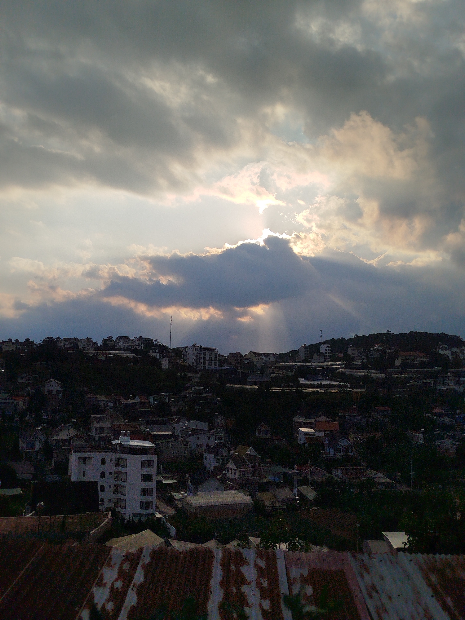 The image shows a cityscape under a dramatic, overcast sky with the sun partially obscured by clouds. Sunbeams break through the cloud cover. The city consists of densely packed buildings, including houses and apartment buildings, situated on a hilly terrain. The foreground features a rusted corrugated metal roof. The location is likely Da Lat, Vietnam. No people are visible.