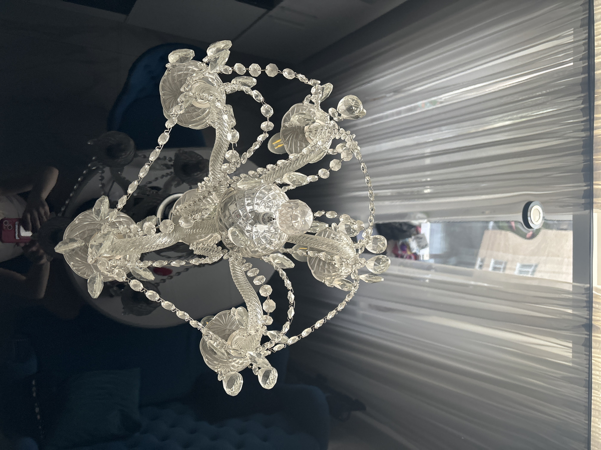 The image depicts a crystal chandelier reflected in a glossy, dark-colored surface, likely a tabletop. The chandelier is ornate, featuring multiple tiers of crystals and glass embellishments. Behind the chandelier, visible through a window or doorway, are sheer white curtains. The reflection also shows a person partially visible in the lower left corner, holding a mobile phone. Beyond the curtains, there is a view of an urban environment with buildings. The location is Unknown, Unknown.