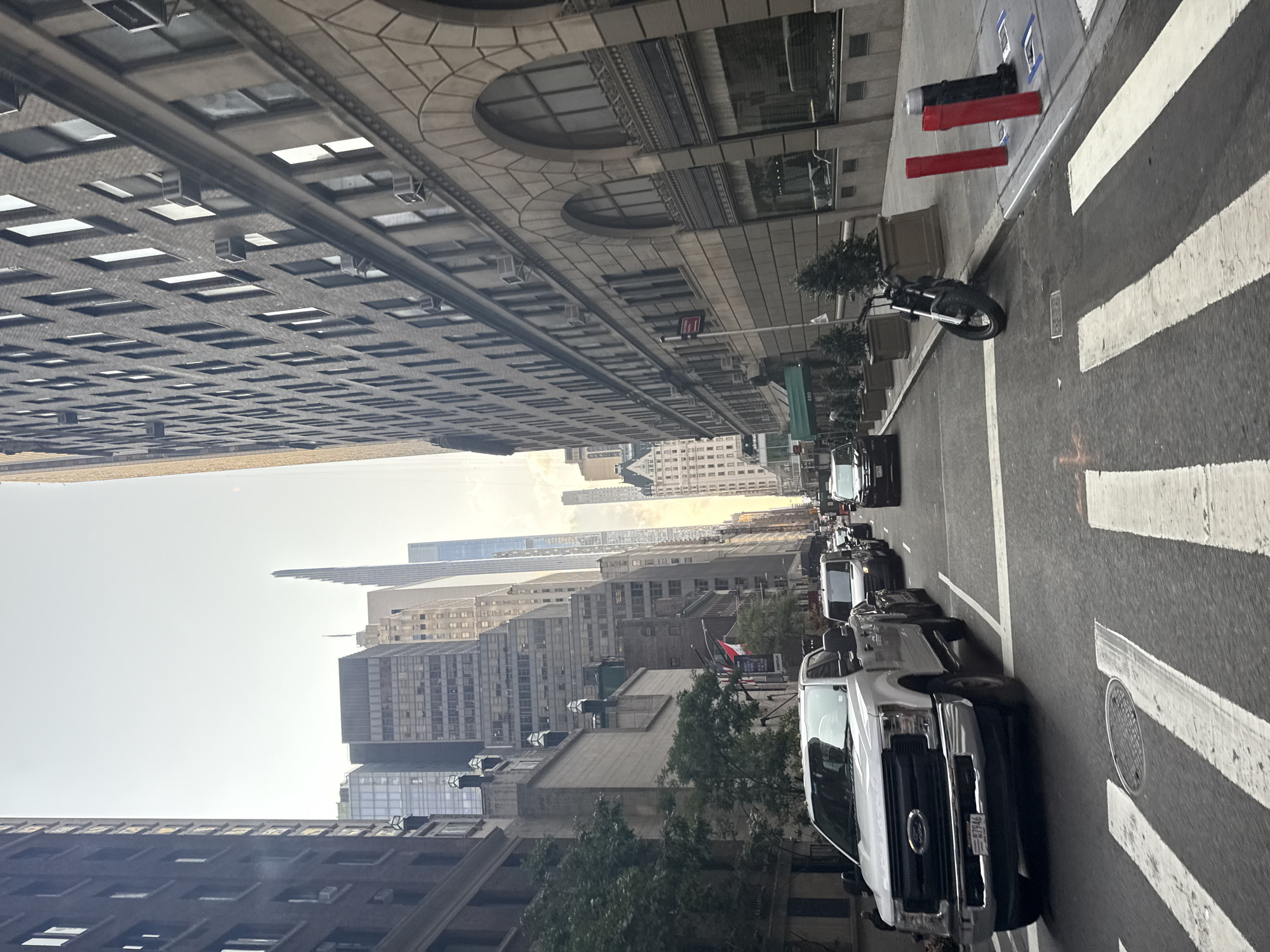 This is a photograph taken from a street in New York City, USA. The perspective is low, looking forward along a street lined with tall buildings on both sides. On the left, a white Ford F-Series pickup truck is in the foreground, partially obscuring the view of other vehicles in traffic. The truck has a license plate that reads "E7946". Behind the pickup truck, there are several other cars and vans in traffic. On the right side of the street, a building with arched windows and ornate stonework is visible. A black motorcycle is parked on the sidewalk next to planters with small trees. Further down the street, numerous other buildings of varying architectural styles and heights are visible, extending into the distance. The sky is overcast. The scene appears to be a typical urban street in Manhattan.