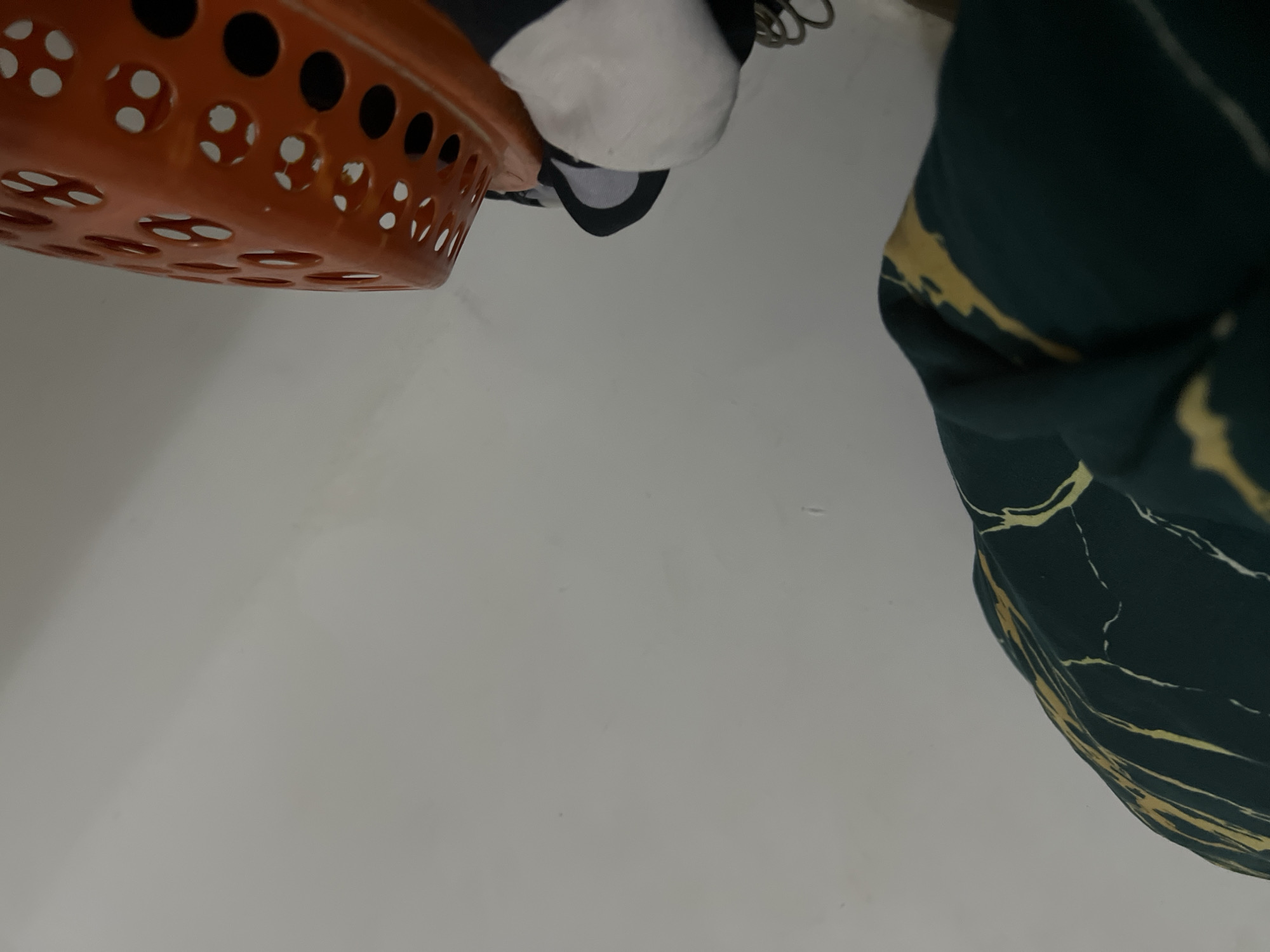 The image shows a close-up view of household items against a plain, light-colored wall. On the left, a dark green fabric with a marbled pattern of yellow and white is draped. To the right, an orange plastic basket with a perforated design is visible. Partially obscured beneath the basket are some white and black fabric items, possibly clothing. The background is a smooth, unadorned wall, suggesting an indoor setting. No specific location in Howayi, Nigeria can be determined from the visual information.