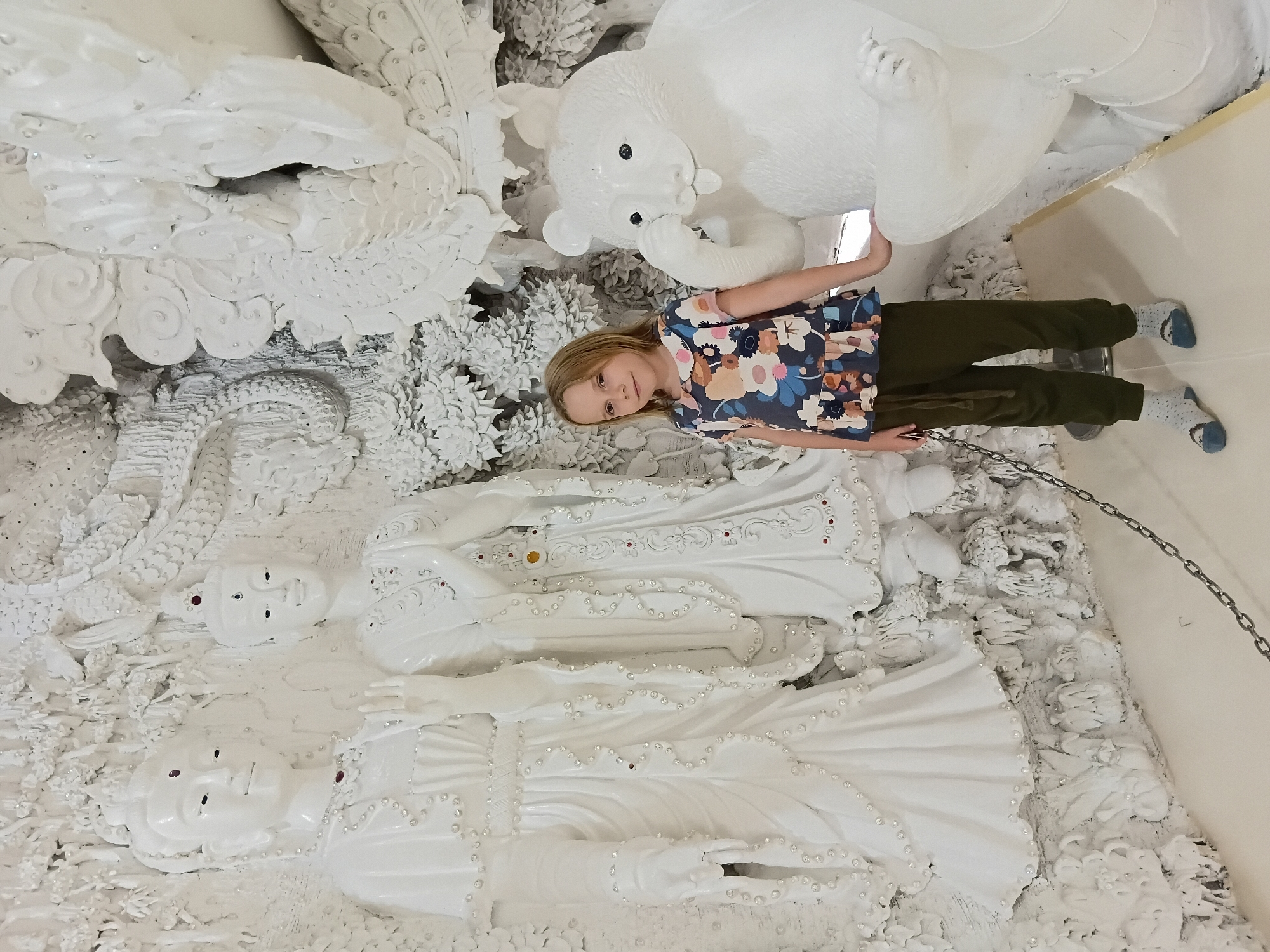 In an indoor setting with white sculpted walls, a young girl stands beside a white statue of a bear. The girl, with blonde hair and wearing a colorful floral-patterned shirt and olive green pants, looks towards the camera. She has one hand resting on the bear statue. The bear statue is depicted with its paw near its nose, and its tongue is slightly out. Behind the girl are two large white statues of human figures, possibly deities or important figures, adorned with small crystals and red gems. The background is an intricate white relief sculpture featuring various motifs. A metal chain is visible in the foreground, extending across the bottom of the frame.