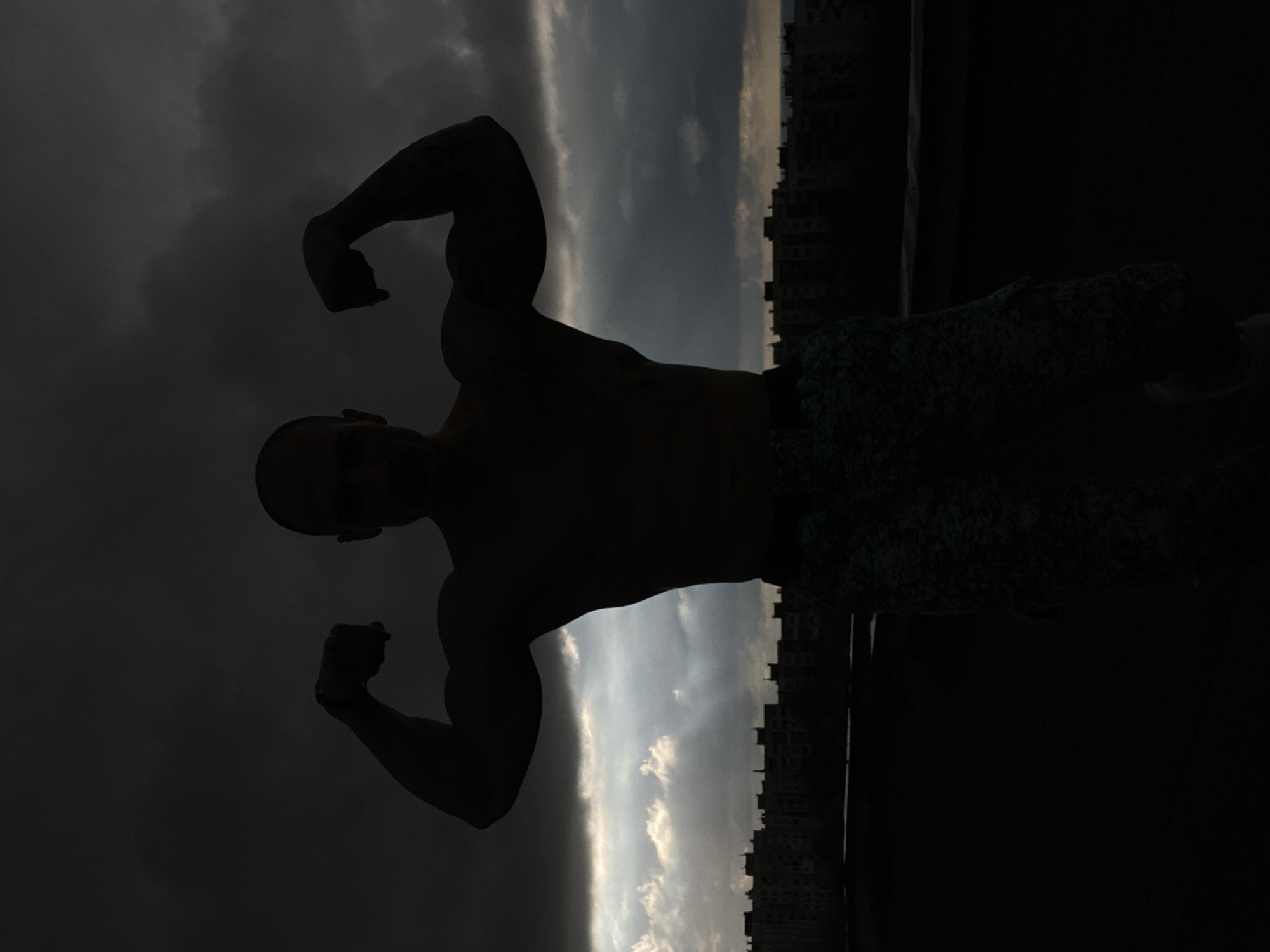 A silhouetted man flexes his arms against a background of a cityscape and cloudy sky. The location is unclear, and there is no way to identify that the image was taken in the city of Pleven, Bulgaria. The man is bald and wearing shorts. The buildings are silhouetted and feature a repetitive window pattern. The sky is partially covered with dark clouds and a strip of lighter clouds.