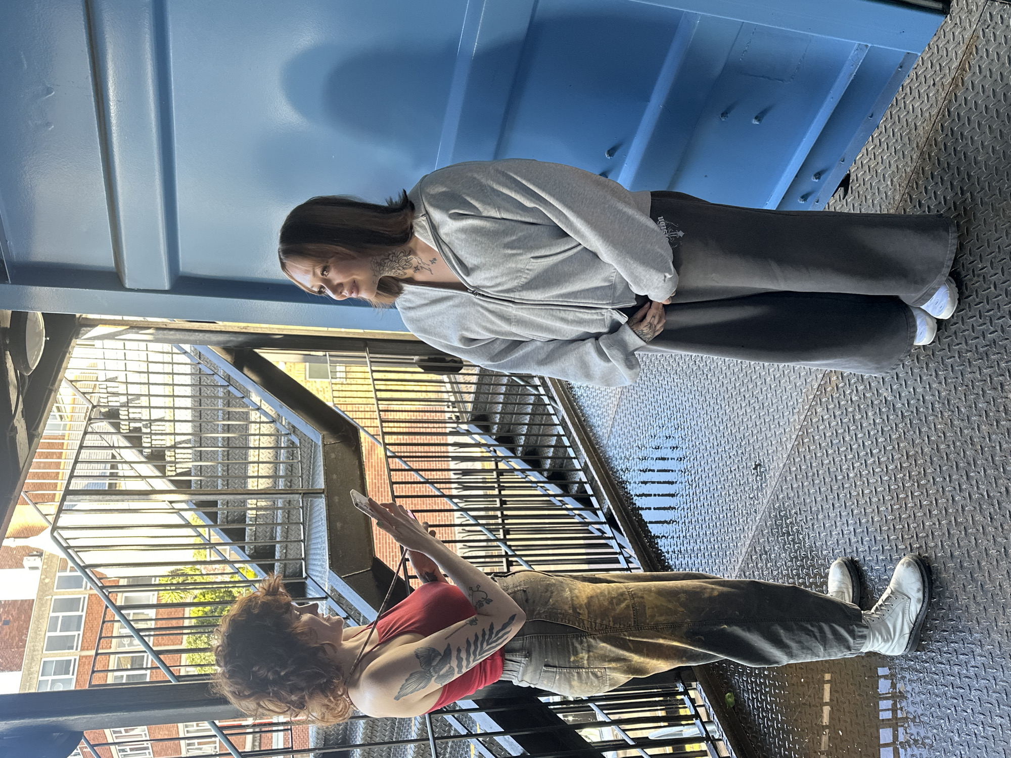 The image shows two women standing on a metal staircase, likely an exterior fire escape, in London. One woman, with curly hair and wearing a red tank top and overalls, is holding a phone and looking at it. The other woman, wearing a grey zip-up hoodie and dark pants, is looking down at the first woman. The environment includes buildings visible through the staircase railings, suggesting an urban setting. The blue metal wall is near the woman in the gray zip-up hoodie. The ground is wet.