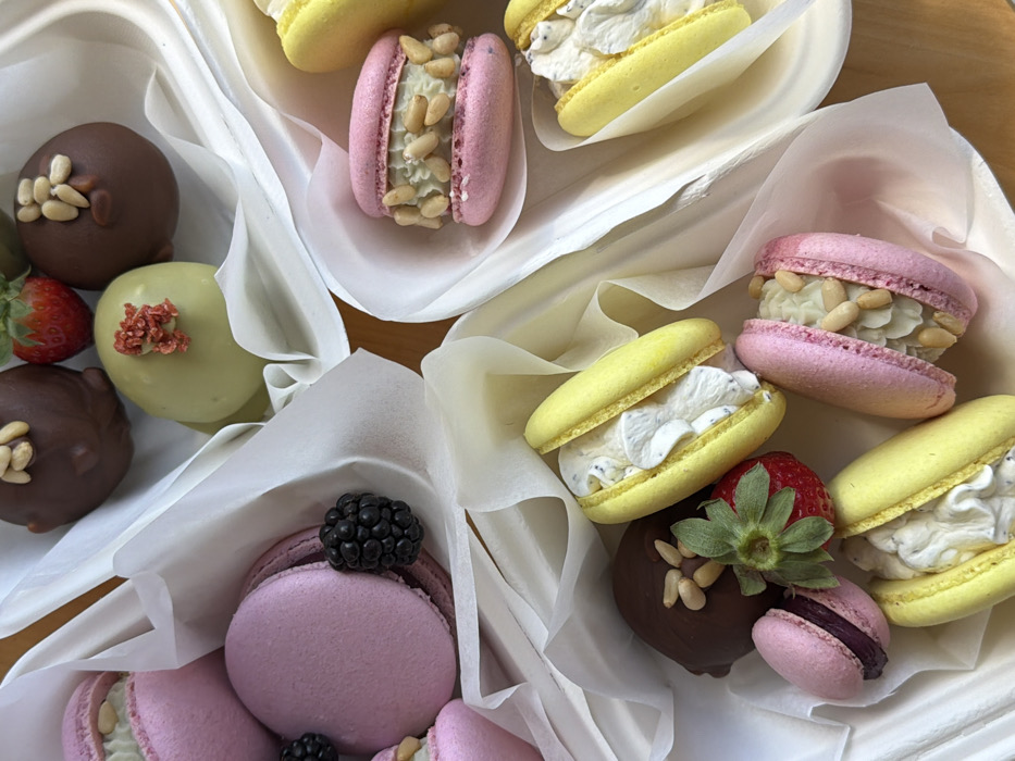 An overhead view depicts an assortment of desserts contained within several white, disposable trays, each lined with white tissue paper. The items are arranged on a light brown wooden surface. The desserts include multiple macarons of various colors: pink macarons, some featuring a creamy white filling topped with pine nuts, others with a darker berry-like filling, and bright yellow macarons, many split open to reveal a white, speckled cream filling. Also present are several spherical, chocolate-covered treats; some are dark brown and topped with pine nuts, while one is light green and garnished with small red dried fruit pieces. Fresh produce includes several bright red strawberries with green stems and one dark blackberry. No specific individuals or actions are depicted. No specific location in Tel-Aviv, Israel, is identifiable from the image content.
