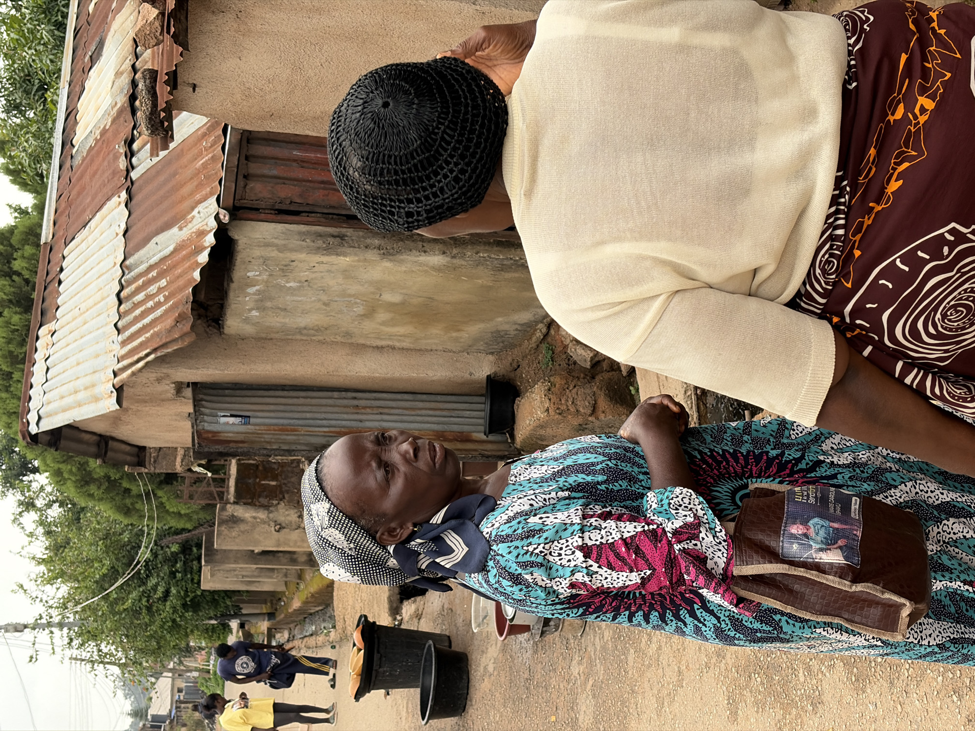Two women are depicted outside a building constructed with rusty, corrugated metal in the city of Jos, Nigeria. One woman is wearing a blue and white patterned headscarf and dress. She is seated with a brown bag on her lap. The other woman, standing, has a black knit cap and wears a cream-colored sweater over a brown patterned skirt. Two people in yellow and blue shirts are walking on the street. Black buckets are in front of the seated woman. The building has a window.