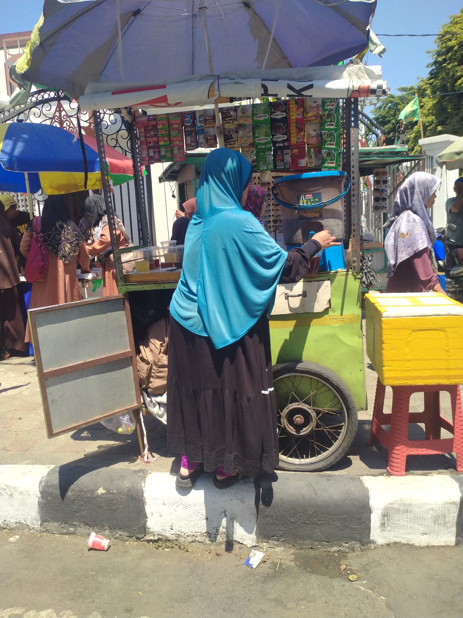 An outdoor street scene in Sukoharjo, Indonesia, features a food cart with a green base and a large blue and white umbrella providing shade. Several individuals, mostly women wearing hijabs, are around the cart. One woman in a turquoise hijab is serving drinks from the cart, holding a small cup. Another woman in a purple hijab stands to the right of the cart, looking towards the front. Behind them, other individuals are partially visible, some interacting with the cart. The cart displays various packaged goods, likely snacks or drinks, on shelves. A yellow cooler sits on a red stool to the right of the cart. The ground is paved, with a raised curb marked with black and white paint. A discarded red cup and other debris are on the ground near the curb. In the background, trees and buildings suggest an urban environment. The lighting indicates it is daytime, with a clear sky.
