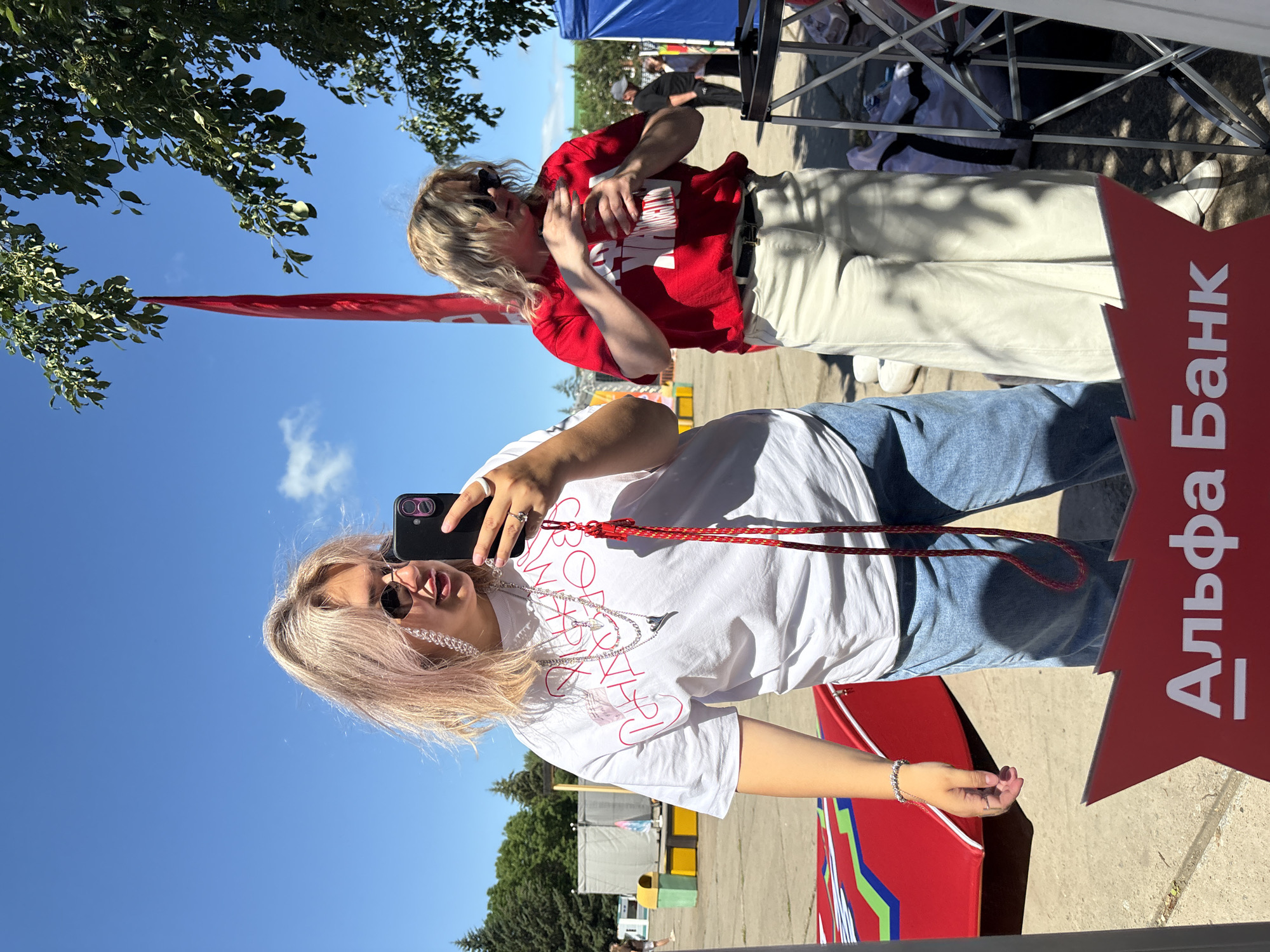 Two young women are standing outside on a sunny day, reflected in a mirror or glass surface. The woman in the foreground, on the left, has blonde hair with pink highlights, is wearing round sunglasses, a white t-shirt with red lettering, and blue jeans. She is holding a black smartphone to take a selfie. She is adorned with several silver necklaces and a silver ring. A red rope hangs from her neck. To her right, partially visible, is another woman with blonde hair, wearing sunglasses, a red t-shirt with white lettering, and light-colored pants. She is holding a cup or bottle. The background shows an outdoor area with trees, a red banner hanging from a tree, and some structures that appear to be part of a temporary setup, possibly for an event. In the foreground, a red object with green and blue accents is visible, and a red sign with white text, "Альфа Банк" (Alpha Bank), is in the bottom center. The overall setting suggests an outdoor public space in Birobidzhan, Russia, possibly during a festival or promotional event.