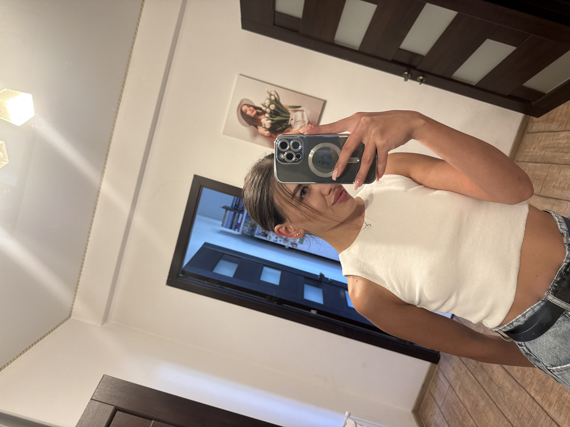 A young woman takes a mirror selfie in what appears to be a residential hallway. She is wearing a white ribbed tank top and denim jeans. In the background, there are two wooden doors, one of which is open revealing shelves with books and other items. A framed picture of a woman holding flowers hangs on the wall between the doors. The ceiling has two light fixtures. The flooring is made of wooden planks. The image does not provide any specific information that can pinpoint the location to the city of Chișinău, Moldova.
