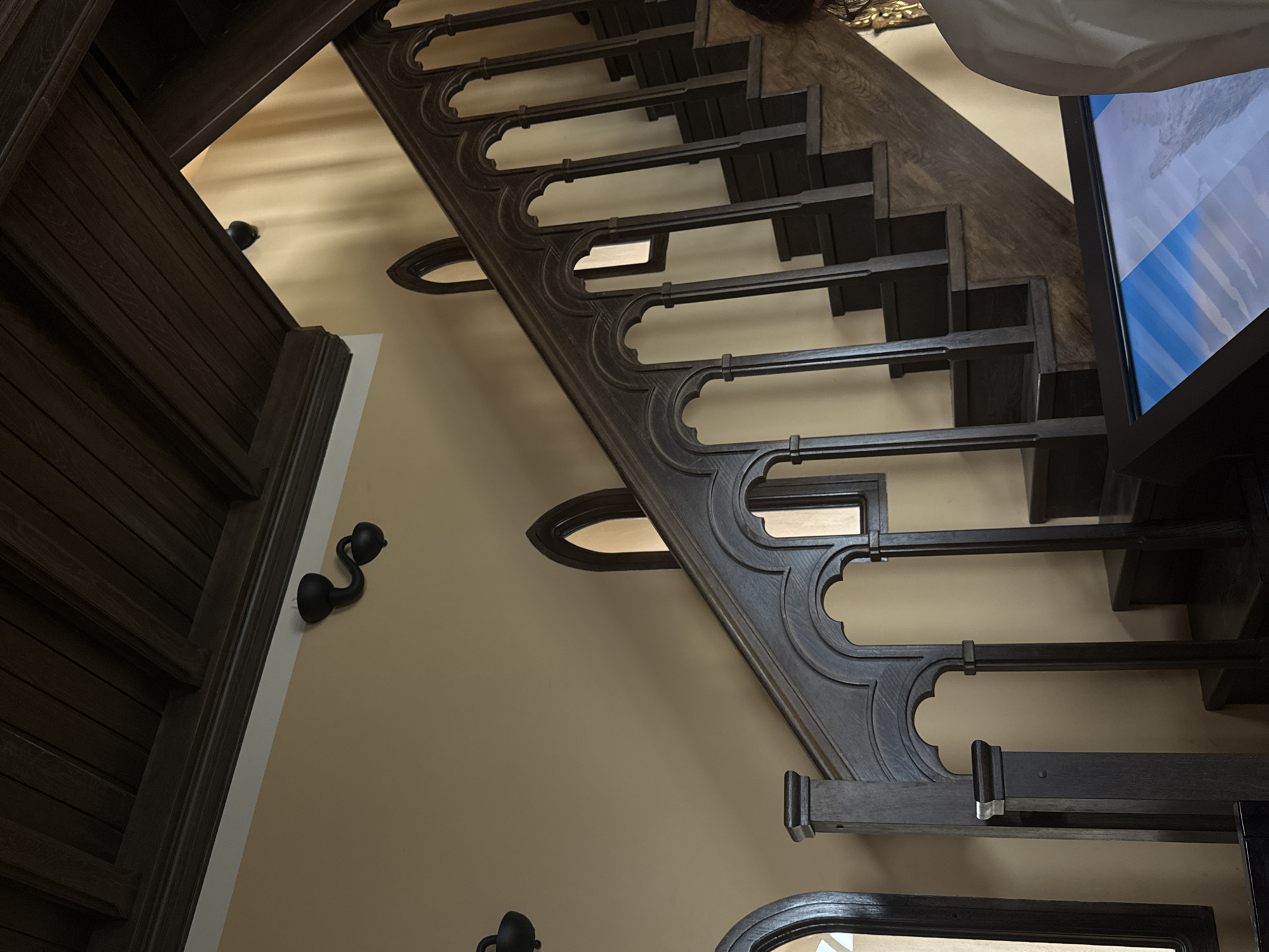 A low-angle shot captures a dark-stained wood staircase with decorative wrought-iron-style railings against a light tan wall. The treads of the stairs are a darker shade of wood. On the left is a section of a wood paneled wall with a sconce, and on the right is a television screen partially visible. The location is Гаспринский поселковый совет, Ukraine.
