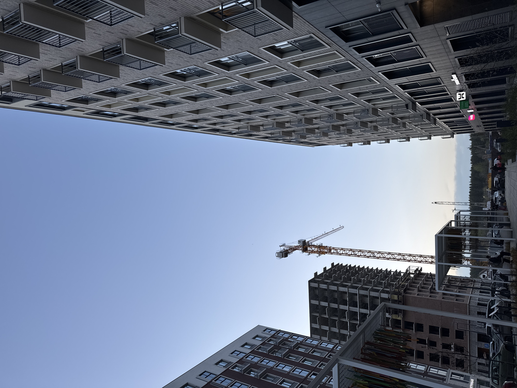 The image presents a view upward in what appears to be an urban environment, likely within the city of Izhevsk, Russia. Two modern buildings are prominently featured on the left side of the frame, their architectural styles contrasting. The taller structure is a light brick building with a grid pattern of windows and external window shades. A construction site is visible on the right side of the image. A crane stands next to a multi-story building that is under construction. In the foreground on the right, there are what appear to be white metal cube art installations on a side-walk. The sky fills a large portion of the frame, transitioning from a deep blue to a lighter shade near the horizon. The photograph is rotated.