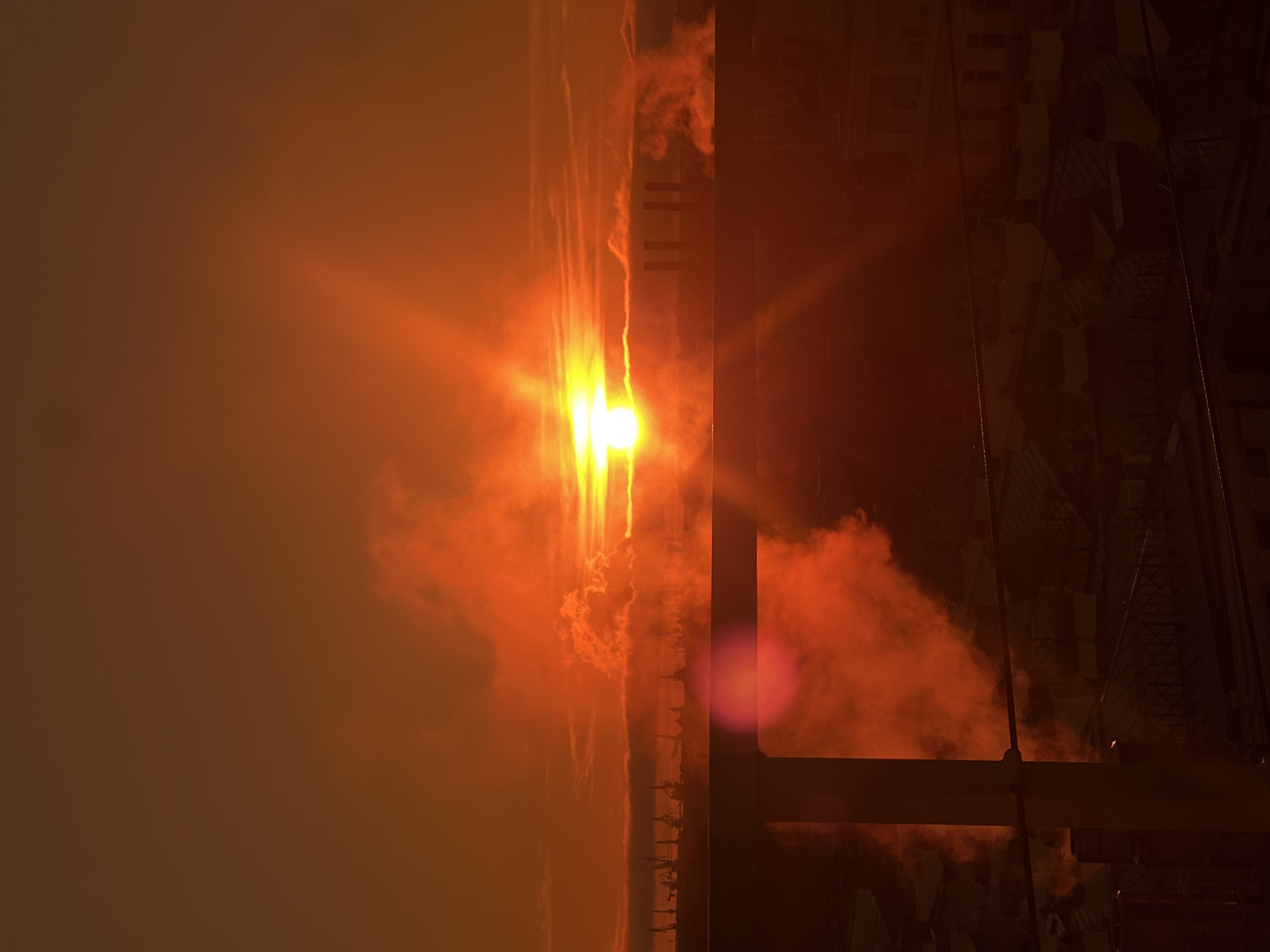 A photograph shows a horizontal view of a fiery sky during a sunset in Saint Petersburg, Russia. The sun, a bright white and yellow orb, is situated near the center, radiating intense orange and yellow light. This light diffuses through what appears to be smoke or fog, casting an overall orange hue across the scene. A series of cranes are silhouetted against the vibrant sky, lined up along the horizon. A building structure partially obscures the view, appearing dark and silhouetted, with its intricate latticework and beams contrasting with the sky.