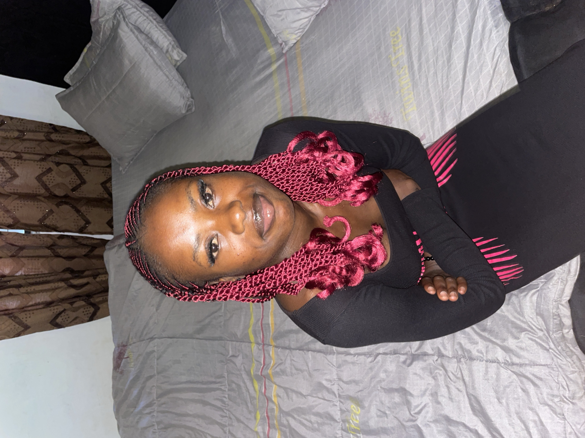 A young Black woman with dark skin and dark hair braided in red twists sits on a bed in a dimly lit room. She is wearing a black long-sleeved dress with pink trim. Her arms are crossed over her chest. The bed is covered in a grey duvet and two grey pillows. Behind the bed, brown patterned curtains hang over a window. The scene appears to be in a residential setting in Jos, Nigeria.