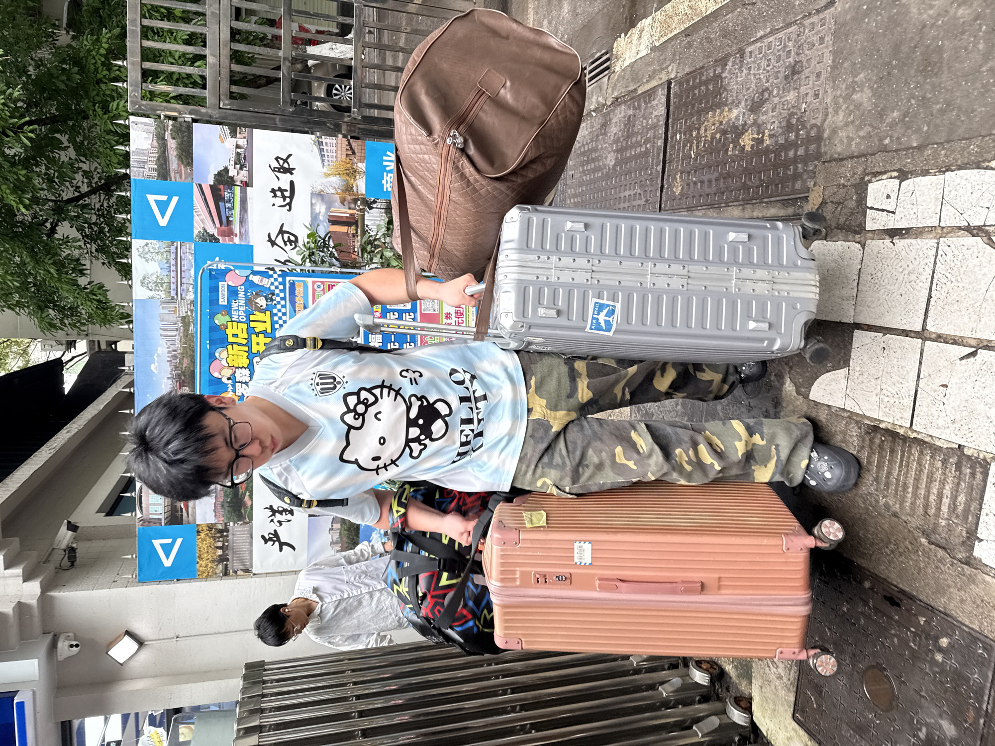 A person wearing glasses and a white and blue shirt with a Hello Kitty design is standing on a sidewalk in the Yuzhong District of China, with three pieces of luggage: a large tan bag, a grey suitcase, and a pink suitcase. A second person in a white shirt and backpack is partially visible. Behind the person are posters advertising a "New Lawson Opening". The sidewalk is made of grey and white tiles, and a metal grate is visible.