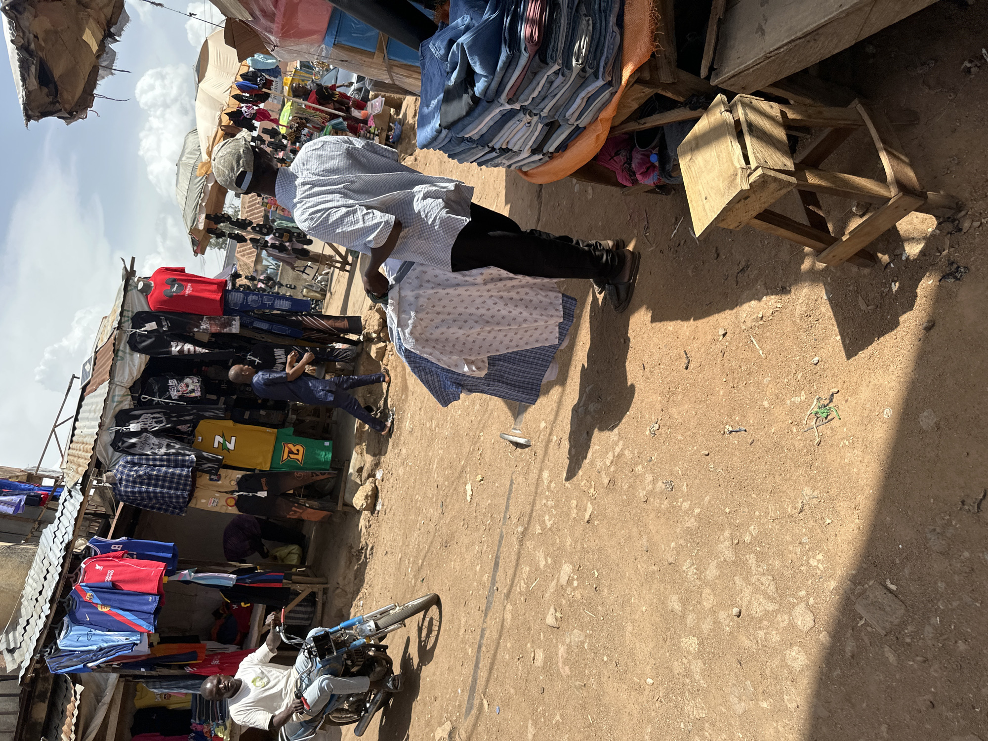 This media file depicts an outdoor market scene, likely in Bukuru, Nigeria. The sun is high in the sky, casting strong shadows, suggesting it is midday. The weather appears to be clear and sunny.
The primary subjects are several individuals, mostly men, engaged in activities related to the market. One man, wearing a light-colored, possibly blue and white checkered shirt and black pants, is standing and looking towards a stall. Another man, in a white shirt and jeans, is seated on a motorcycle, looking towards the viewer. Other individuals are visible in the background, appearing to browse or work at the stalls.
The setting is a bustling marketplace with numerous stalls selling clothing. Clothing items, including t-shirts, shirts, and possibly pants, are displayed on racks and shelves. There are also stacked garments, likely jeans, visible on a wooden structure. Several umbrellas and awnings provide shade. The ground is dusty and uneven, typical of a market in this region.
Objects of note include a motorcycle, wooden crates and structures used for display, and various articles of clothing. There is also a pile of blue fabric at the top of the image.
The overall mood seems to be one of everyday commerce. The individuals appear focused on their tasks or interactions within the market environment.
Visible text is minimal, but some clothing items have logos or designs that might be brands or team insignias. For instance, a red t-shirt with a white Mickey Mouse logo is visible, and a yellow t-shirt with the letter "N" and the number "27" is also present.