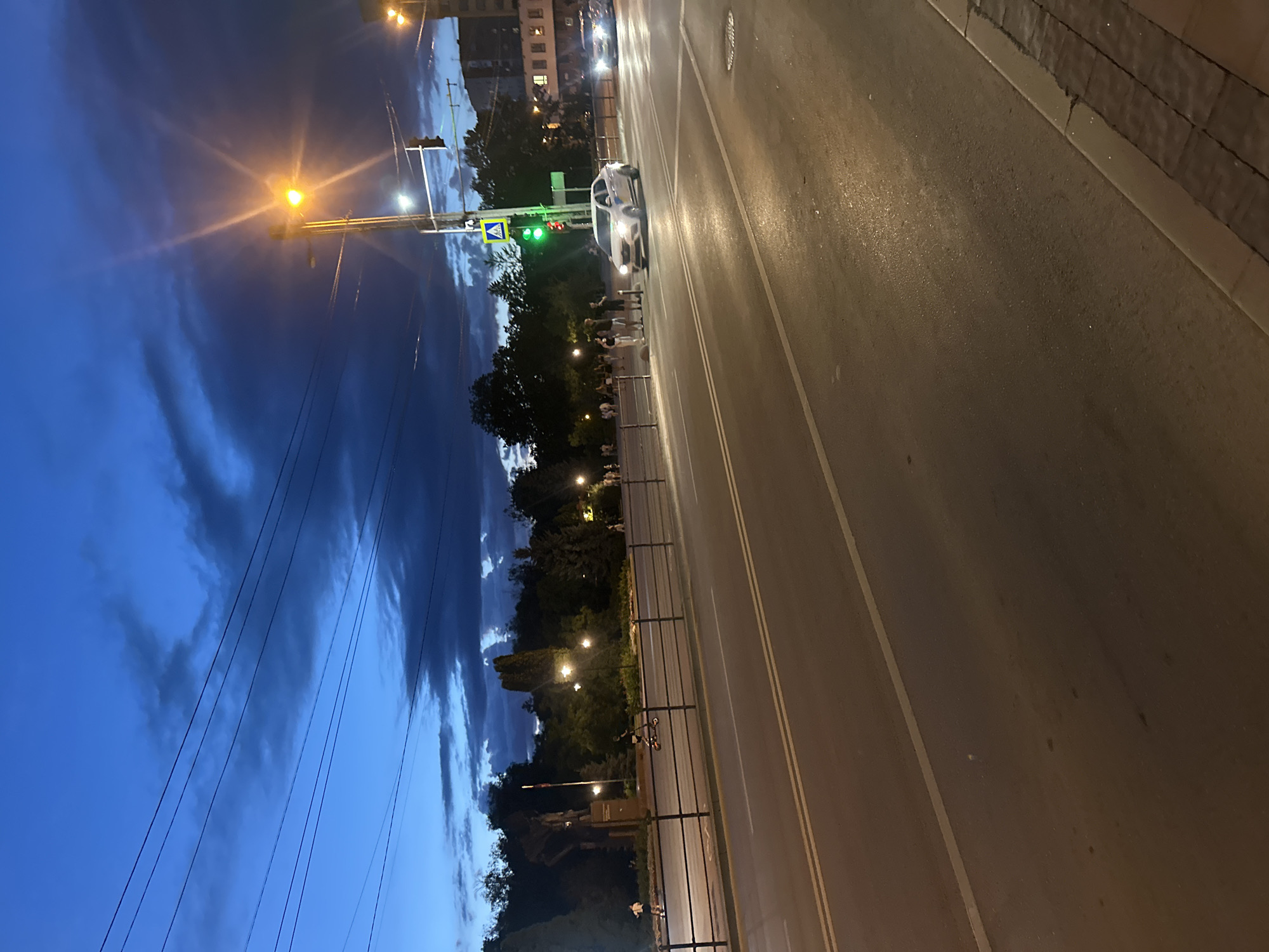 The image captures a street scene in Ternopil, Ukraine, at dusk. The sky is a deep blue with dramatic dark clouds, illuminated by a bright street lamp emitting a yellowish glow, creating a starburst effect. Multiple power lines crisscross the sky.
The foreground shows a wet asphalt road with yellow lane markings. To the left, a black metal fence separates the road from a pedestrian walkway and a park-like area. On the walkway, several people are visible. Some are standing in a group near a crosswalk, while others are scattered further along. A person is riding an electric scooter in the middle ground. In the background, a statue or monument is partially visible amidst trees.
A white car is driving across the crosswalk. Traffic lights are visible, with a green light illuminated for the cars. Further down the street, another car with headlights on is approaching. The overall atmosphere is that of a city street transitioning from day to night.