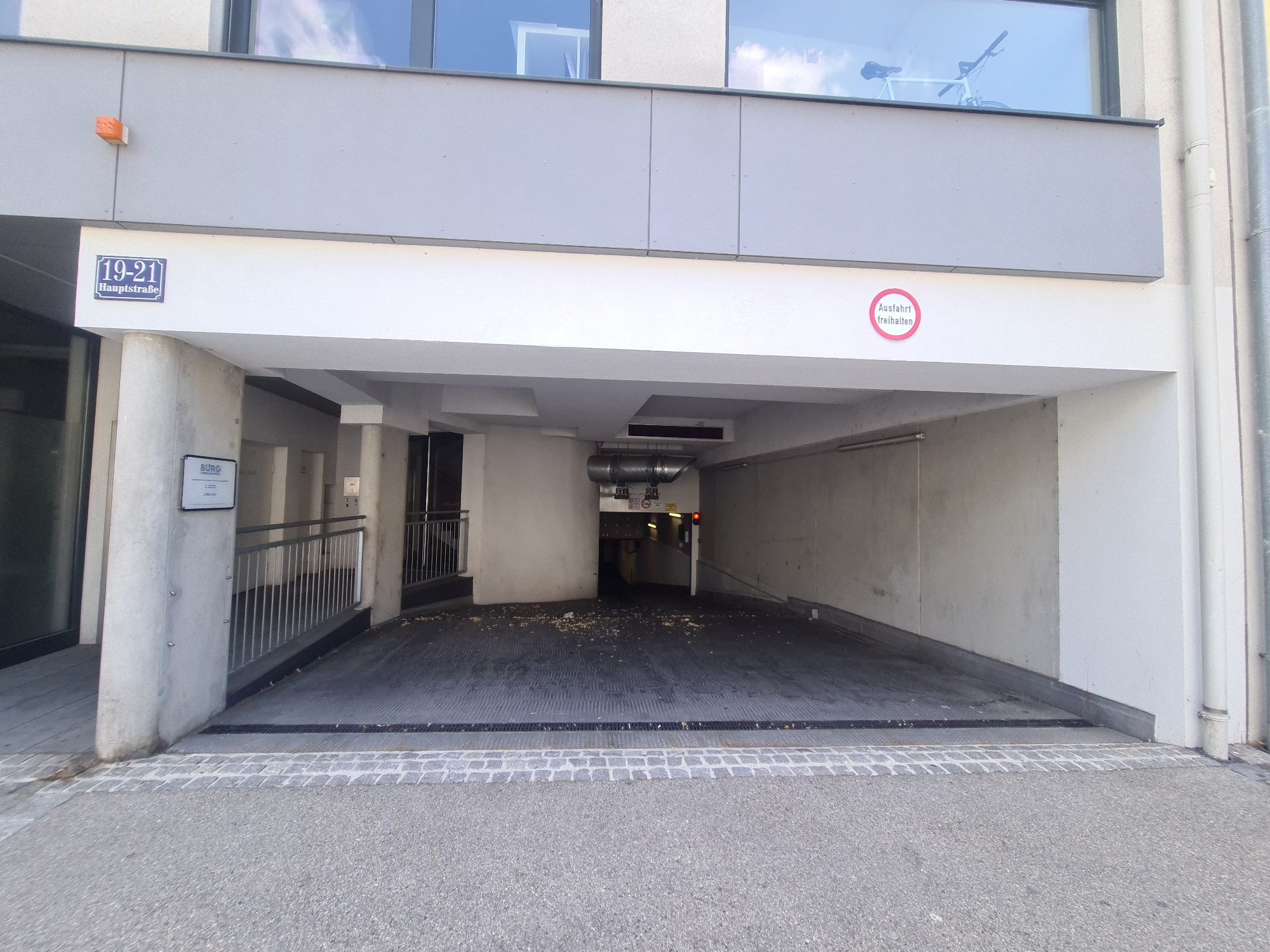 The image shows the entrance to a parking garage. A sign with "19-21 Hauptstraße" is visible on the wall above the entrance, indicating the address. A red circular sign with "Ausfahrt freihalten" is also visible, meaning "Keep exit clear." The parking garage has a ramp leading down into it, and the ground appears to be wet in places. There are some debris on the floor of the garage. The lighting suggests it is daytime, and the weather appears to be overcast. The scene is an urban setting, likely in Mödling, Austria. There are no people visible in the image. A bicycle is visible through a window on the upper floor of the building.