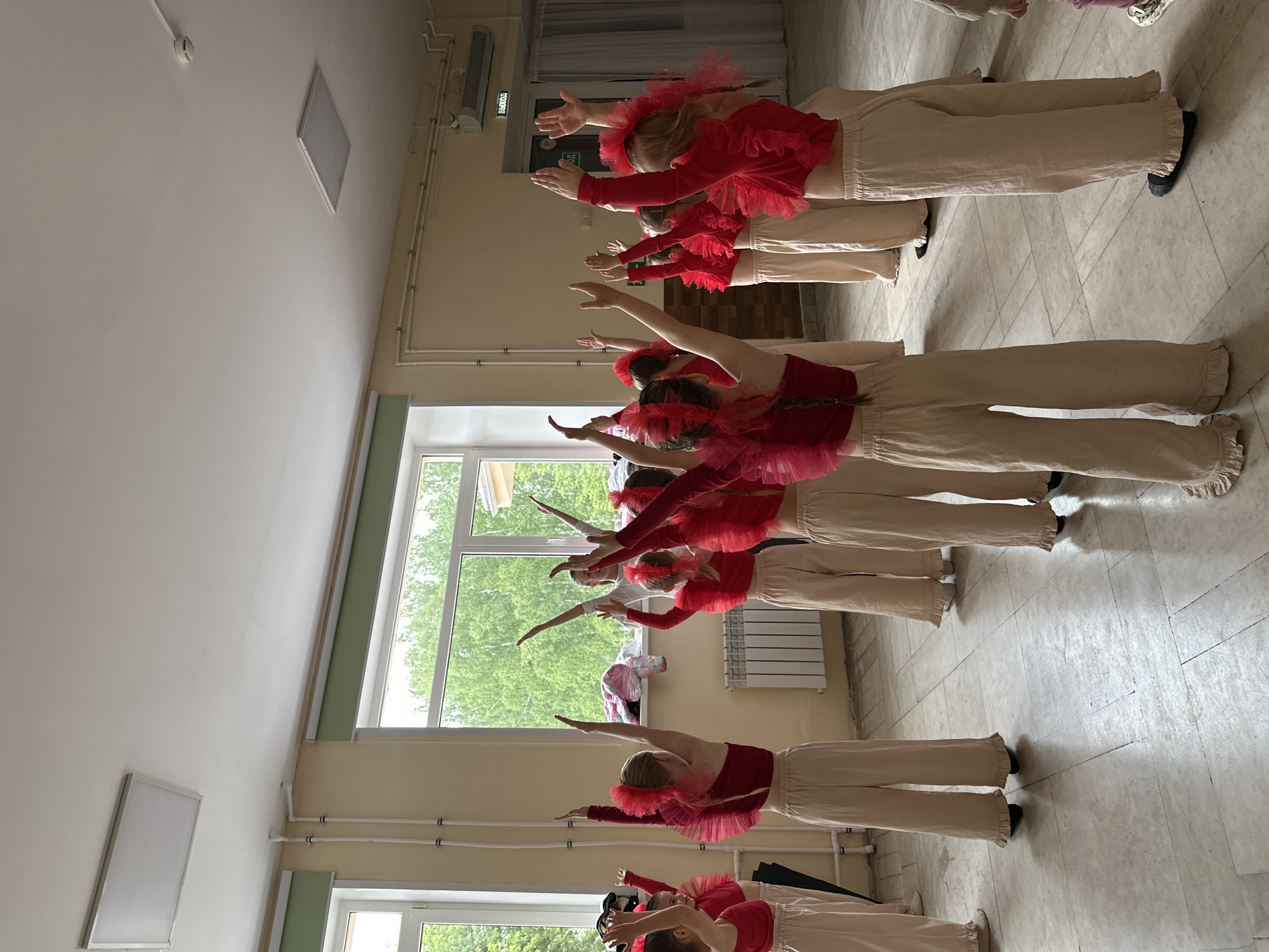 The image captures an indoor scene featuring a group of young children, predominantly girls, performing a synchronized dance or exercise routine. At least seven children are visible, arranged in rows or lines, facing towards the right side of the frame. They are uniformly dressed in vibrant red tops, which feature ruffled or tulle accents around the shoulders, neckline, or waist/hips. These are paired with loose-fitting, light beige or tan pants, some with ruffled cuffs at the ankles, and black shoes. Several children have their hair tied back, often adorned with red accessories matching their outfits.
All participants are performing a coordinated pose: their arms are extended forward and slightly upward, with open hands. They appear focused and engaged in the activity.
The setting is a brightly lit room, likely a dance studio or multi-purpose hall. The walls are a light cream or off-white color, meeting a white ceiling where exposed white pipes or conduits run alongside various light fixtures, including a rectangular panel and a smaller circular one. Large windows along one wall allow significant natural light to enter, revealing lush green foliage outside, indicating a daytime scene during a warm season. A white radiator is visible beneath one of the windows. The floor is covered with light-colored, square tiles.
In the background, above a doorway, a red emergency exit sign is prominent, displaying the Russian word "ВЫХОД" (VYKHOD), confirming the specified location context of Chaikovsky, Russia. A plain white rectangular board is also mounted on a wall in the upper-left background. The overall impression is one of an organized and well-lit space for children's activities.
