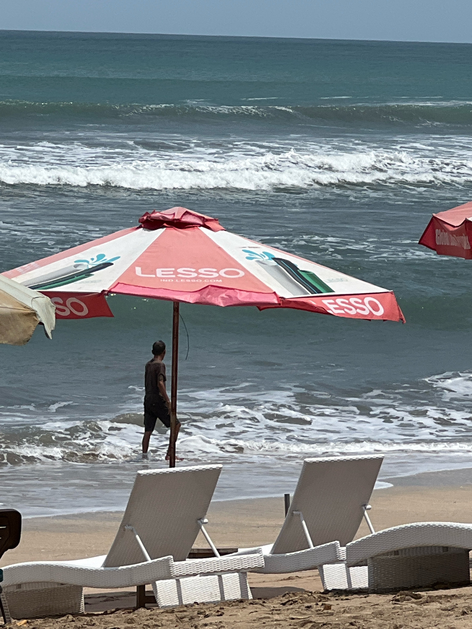 The image depicts a beach scene in Kuta, Indonesia. In the foreground, two white sun loungers are positioned on the sand. Behind them, a man is standing in the shallow water, partially obscured by a large pink and white umbrella. The umbrella has the "LESSO" logo printed on it. The ocean is visible in the background with moderate waves. Another red umbrella is partially visible to the right. The sky is clear and blue. The overall impression is a relaxed beach setting on a sunny day.