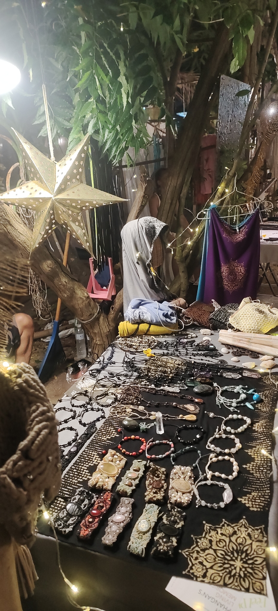 This image depicts an outdoor market stall at night, likely in the Ko Pha-ngan District, Thailand, given the presence of what appears to be a "Phangan's Living Memories" tag. The stall is laden with various handmade jewelry and crafts, predominantly necklaces and bracelets made of beads, stones, and macrame. A large, illuminated star decoration hangs prominently from a tree branch. In the background, a person is partially visible, dressed in a grey hooded garment, and appears to be attending to the stall. Several fabrics, including a purple and blue patterned textile, are displayed on hangers. The ground appears to be dirt or sand, characteristic of an outdoor setting in Thailand. String lights are draped around the stall, illuminating the merchandise and creating a festive atmosphere. A pink child's chair is also visible near the tree.