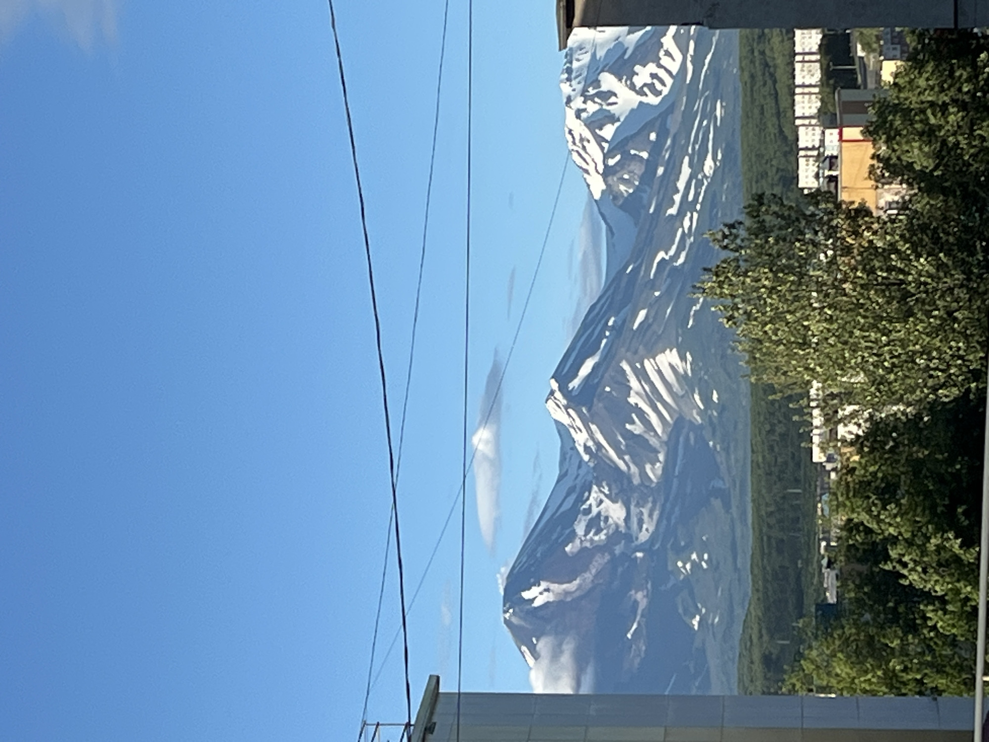 A bright, clear blue sky dominates the upper portion of the image, with three distinct power lines stretching horizontally across the frame. Below the sky, a range of majestic, snow-capped mountains rises. The largest mountain in the foreground, with a conical peak, shows patches of snow accentuating its slopes. To its right, another substantial snow-covered mountain is visible, with a flatter, plateau-like summit. Further to the right, a third snow-capped peak completes the mountain panorama. The mountains are partially obscured by wispy white clouds clinging to their upper reaches. A lush green forest forms a dense band across the mid-ground, separating the mountains from the urban landscape below. In the foreground and mid-ground, parts of buildings and trees are visible. The left side of the image is framed by the corner of a light-colored building, and the right side is partially obstructed by another building. The scene is likely Petropavlovsk-Kamchatsky, Russia, given the characteristic views of the surrounding volcanoes.