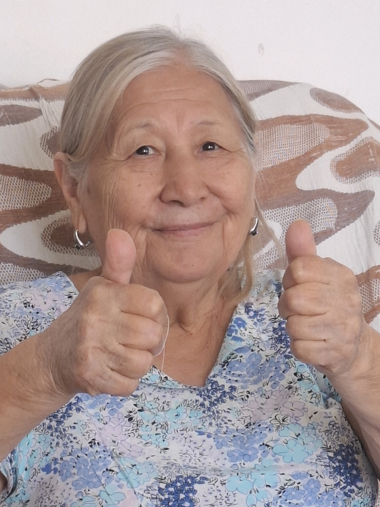 An elderly East Asian woman with grey hair is shown from the chest up, sitting in a chair with a patterned cushion behind her. She is smiling and giving a thumbs up with both hands. She is wearing a blue and white floral print blouse and small hoop earrings. The background is a plain white wall. The image was taken in Almaty, Kazakhstan.