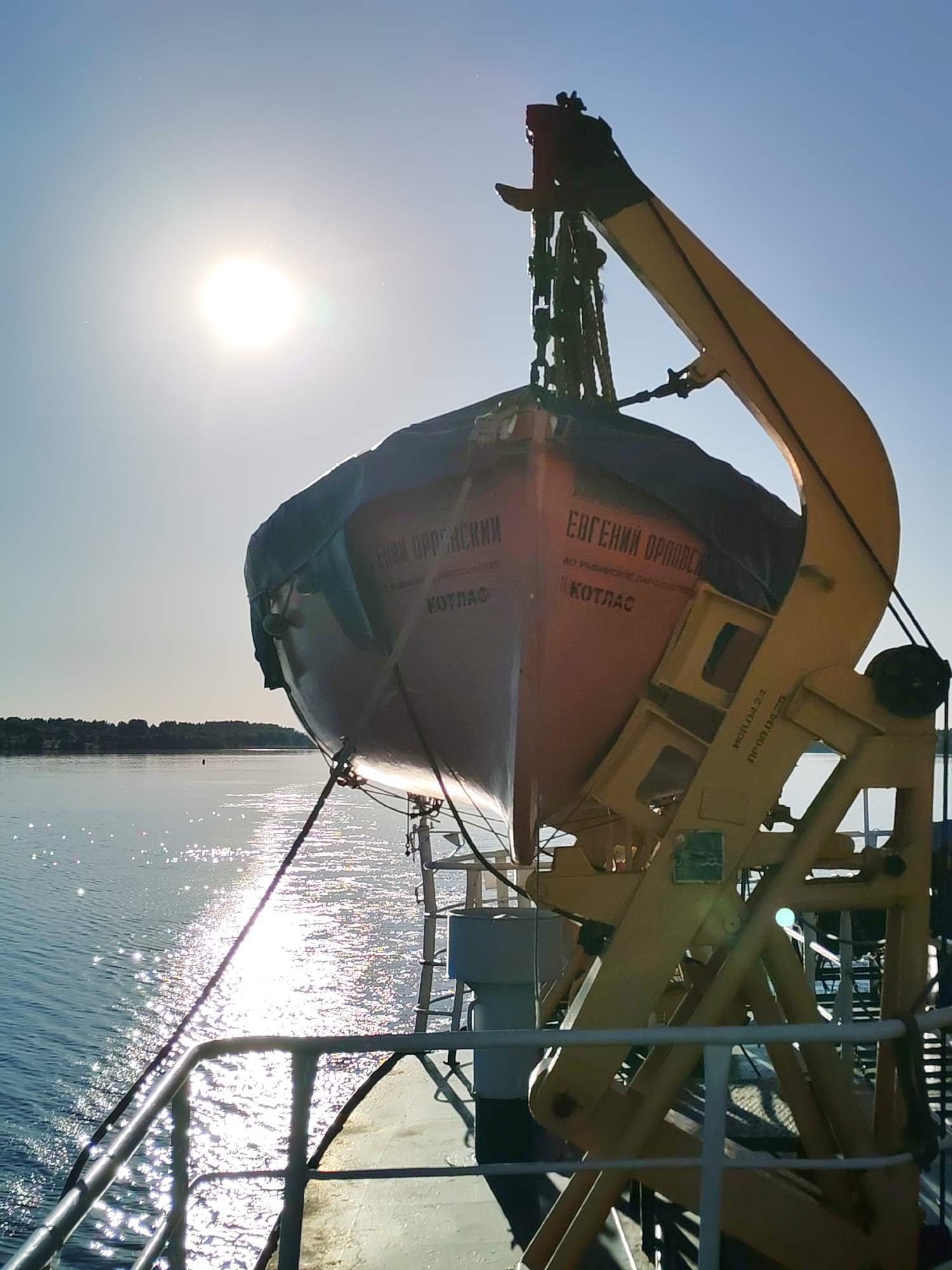 This is a photographic image depicting a maritime scene, observed from the deck of a vessel. The primary subject is an orange-red lifeboat, partially covered with a dark blue or green tarpaulin, suspended from a robust yellow davit system on the starboard side of the ship. The lifeboat's hull displays Cyrillic text: "ЕВГЕНИЙ ОРЛОВСКИЙ" (YEVGENIY ORLOVSKIY), "АО РЫБФЛОТ-РК" (AO RYBFLOT-RK, a Joint Stock Company "Fish Fleet-RK"), and "КОТЛАС" (KOTLAS). The yellow davit system, which secures the lifeboat above the water, also bears stenciled markings, including "ИЭП.04.24" and "ПР.00.04-2Б". Ropes and chains are visible connecting the lifeboat to the davit.
The foreground features a white or light grey ship's deck and a metal railing. Beyond the ship, a wide body of calm water stretches towards the horizon. The surface of the water reflects intense sunlight from the upper left, creating numerous bright, shimmering highlights. In the distance across the water, a low-lying landmass covered with a treeline is discernible. The sky is clear and light blue, with a very bright, circular sun positioned in the upper left quadrant, generating lens flare. No individuals are present in the frame. The scene is illuminated by direct, strong daylight, creating contrasting areas of light and shadow on the equipment. The location is observed from a vessel in the city of Mys, Russia, with the lifeboat indicating "Kotlas" as an associated port.