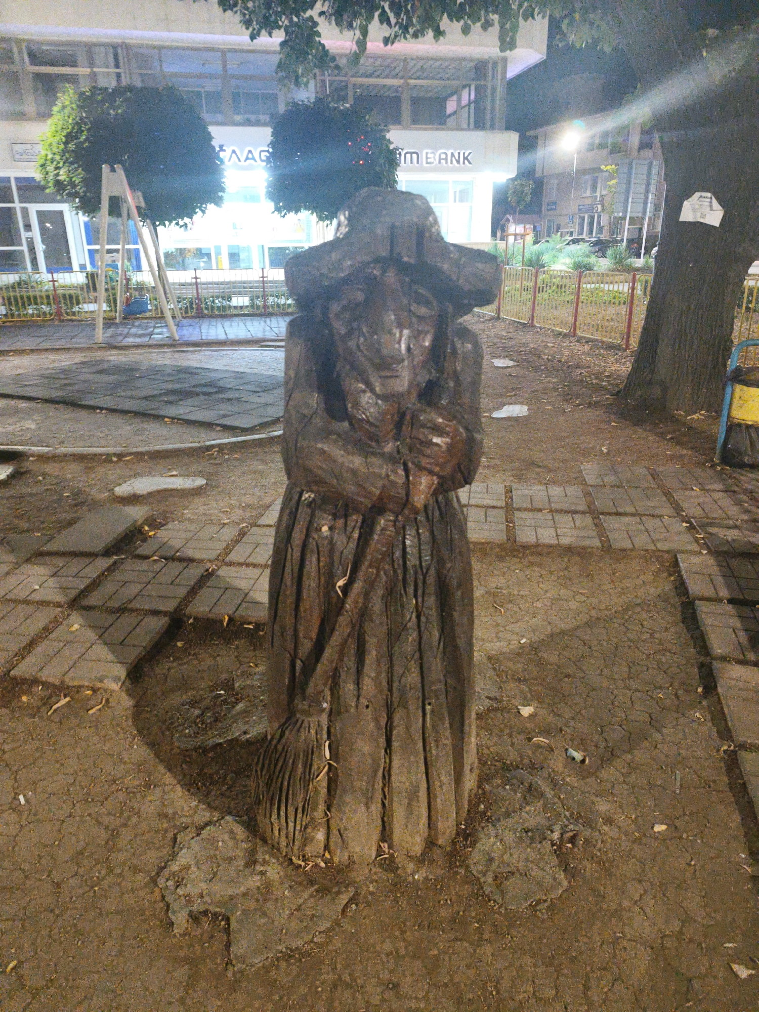 The image shows a nighttime exterior scene in the city of Lovech, Bulgaria. In the foreground, a wooden sculpture of a witch holding a broom stands centrally. The sculpture appears weathered and is made from a dark wood. It is positioned on a paved area with some fallen leaves and dirt. Behind the sculpture, a paved pathway leads away from the viewer, bordered by a yellow and red fence. To the left of the sculpture, a playground area with rubber matting and a swing set is visible. In the background, there are buildings, including one with signage that appears to read "MAAG" and "M BANK." Streetlights illuminate the scene, casting shadows and highlighting the textures of the objects. A large tree trunk is present to the right of the sculpture, and a trash bin is visible near the tree. The overall impression is of an outdoor public space at night, with the unique wooden sculpture being the primary subject.