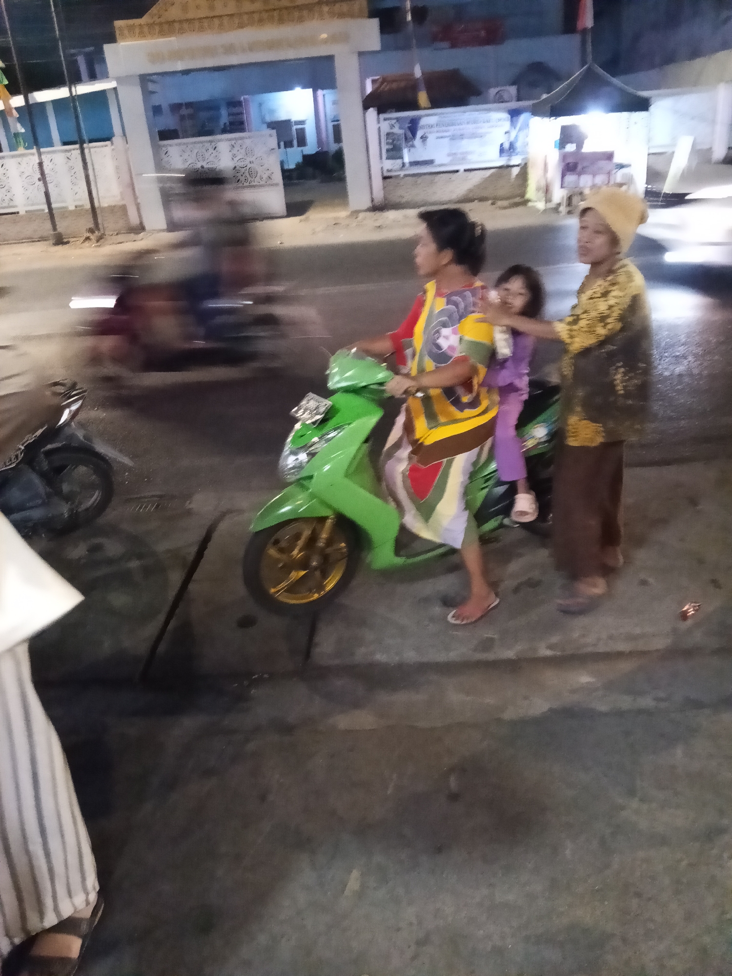 In the city of Lubuklinggau, Indonesia, three individuals are depicted on a street at night. A woman in a brightly colored shirt and skirt is operating a green scooter, with a young child seated behind her, wearing a purple outfit and pink sandals. A third individual, wearing a yellow patterned shirt and brown pants, stands beside the scooter, also appearing to be on the street. The environment suggests a commercial or residential area with a building and some tents in the background, illuminated by artificial lighting. The scooter's wheels are visible, with one featuring gold-colored rims. Another dark-colored motorcycle is parked to the left of the scene. The image is slightly blurred due to motion or low light conditions.