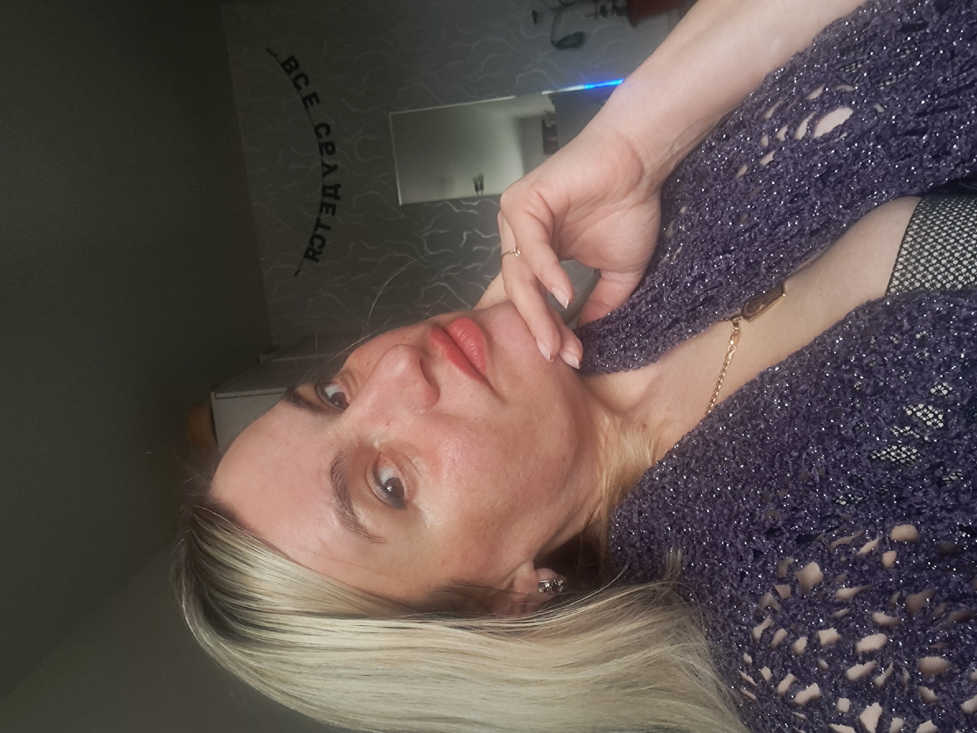A close-up portrait shows a woman with long, straight blonde hair, possibly highlighted, looking directly at the camera. Her lips are painted a reddish-pink, and her expression is neutral to contemplative. She wears a dark purple, open-knit top or cardigan with a glittery finish, and a gold chain necklace is visible. A hint of a black, fishnet-style garment is visible underneath her top on the left side of the frame. Her right hand, adorned with a small ring on the ring finger, is resting on her chin and jawline.
The setting is indoors, with a patterned wall in the background. A rectangular mirror is visible on the wall, reflecting a portion of the room. Above the mirror, Russian text is visible on the wall, which reads "ВСЕ СРАЗУ ДЕЛАЮ" (VSE SRAZU DELAYU), translating to "I DO EVERYTHING AT ONCE." A small blue light source is also present in the upper background, possibly from an electronic device. The image is oriented with the woman's head tilted significantly to the right, suggesting it was originally taken in portrait mode and then rotated, or the subject herself was lying down. The overall ambiance is personal and direct. This scene is located in Elektrostal, Russia.