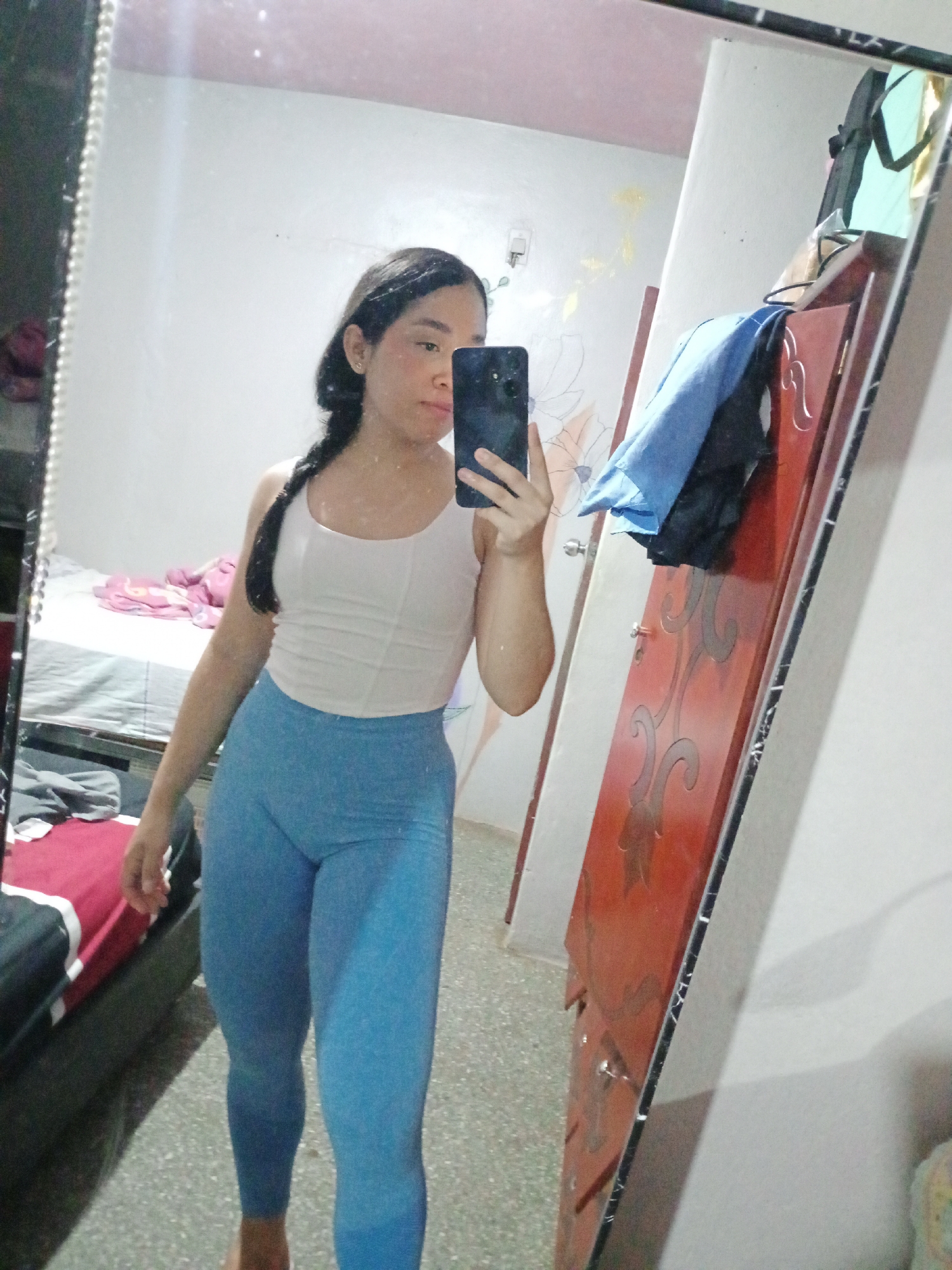 A woman is taking a mirror selfie in what appears to be a bedroom. She is wearing a light-colored top and blue leggings. She is holding a smartphone in her right hand, partially obscuring her face. Her hair is dark and styled in a braid. The bedroom contains a bed with a red and grey patterned comforter and a mirror in which the woman is reflected. A wooden door is visible in the background. It is not possible to determine the location in Valle de La Pascua, Venezuela, from the image.