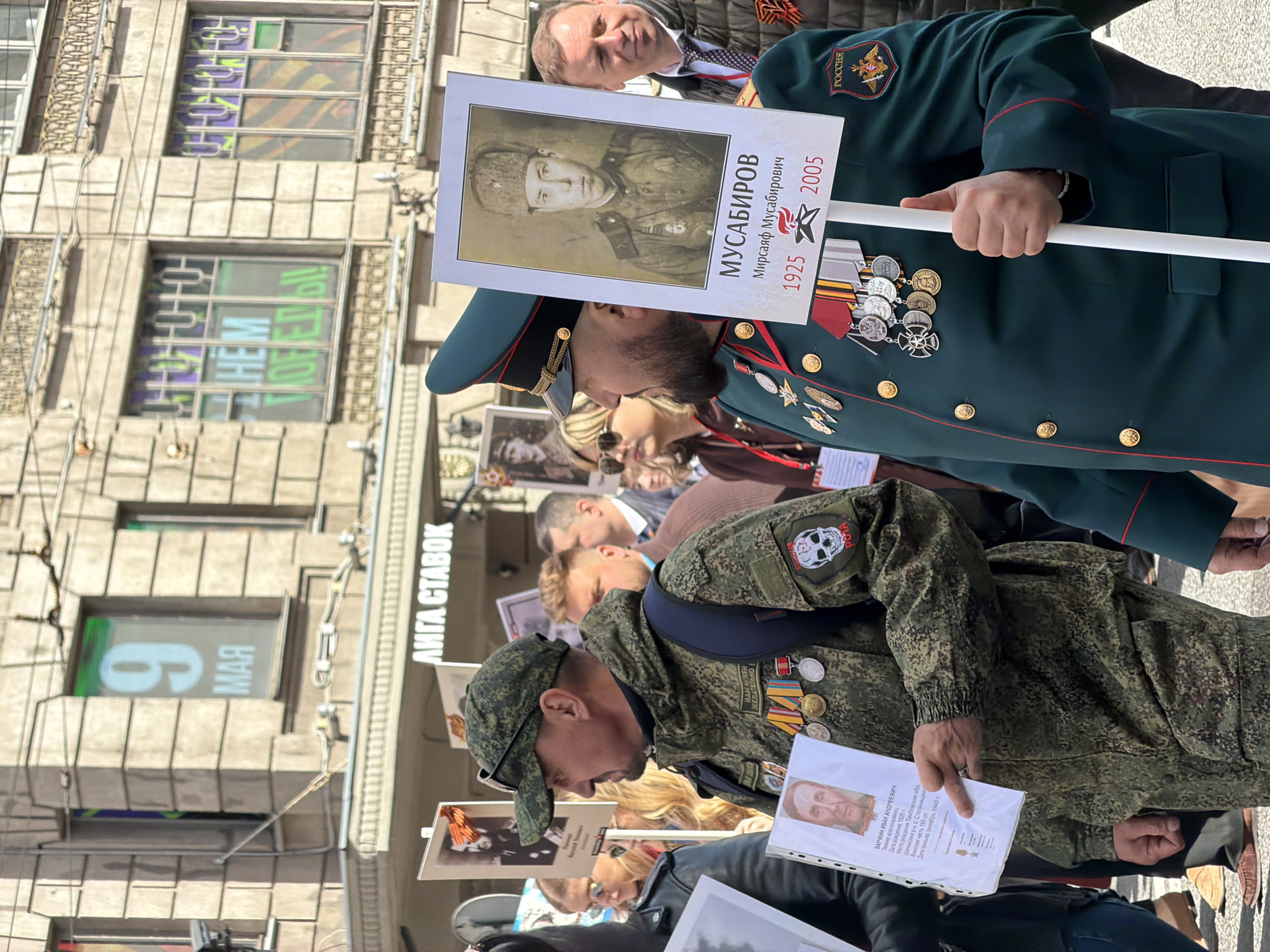 The image shows a group of people in what appears to be a street parade or memorial event in Saint Petersburg, Russia. Several people are holding portraits of individuals, likely veterans or those who died during wartime. One man in a green military uniform is holding a sign with a photo and the name "МУСАБИРОВ Мирсаяф Мусабирович" along with the years "1925-2005" and a symbol resembling a star. Another person in a camouflage military uniform is holding a document with a portrait and text, including the name "БАУЧКИН ИВАН АНДРЕЕВИЧ" and information about his birth year (1920) and military service. Both men are wearing military decorations. A building is visible in the background. The windows have signs that read "HEM ГЛОБЕДЫ!" and "9 МАЯ." Part of a larger sign reads "ЛИГА СТАВОК." The attire and signs suggest that the event is related to Victory Day, commemorating the end of World War II.