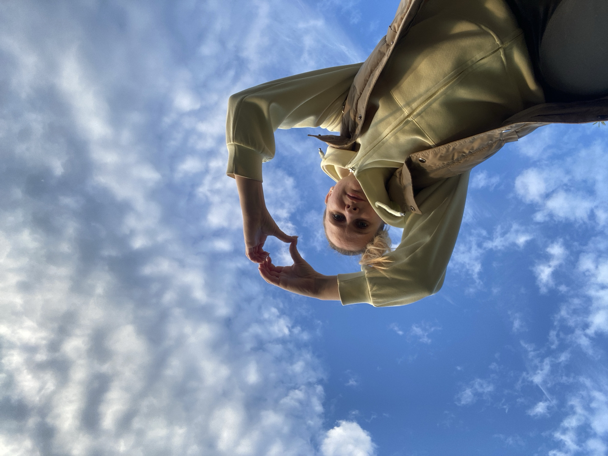 A young woman is making a heart shape with her hands, taken from a low angle against a blue sky with scattered clouds. She is wearing a light yellow hoodie with a brown vest. Her hair is pulled back. The location is Chaikovsky, Russia.