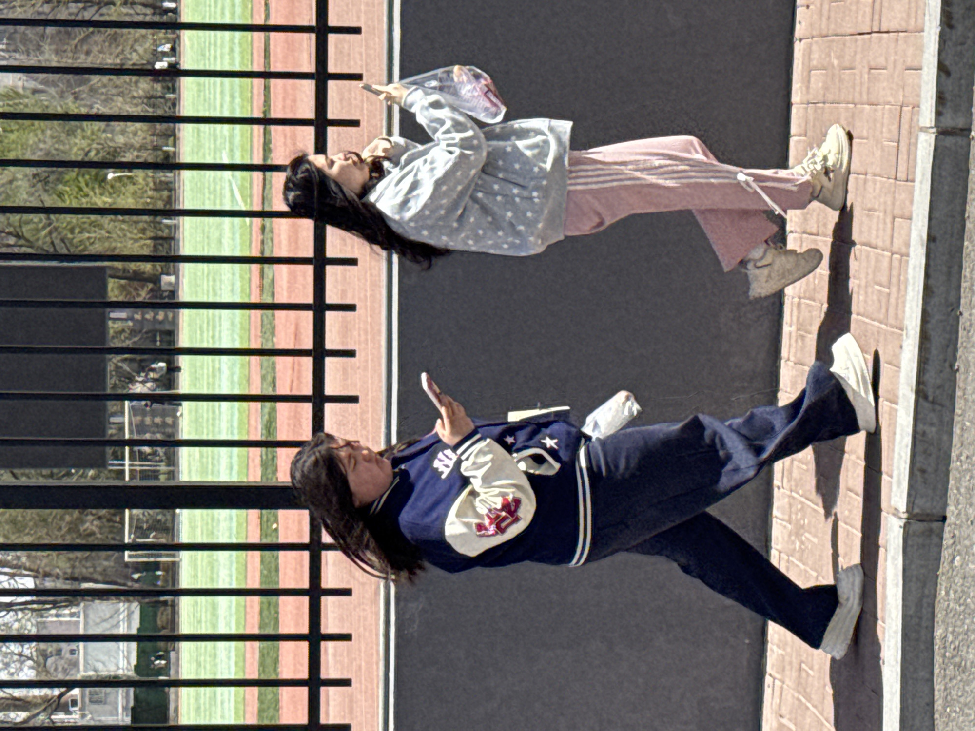 Two East Asian women stand on a brick sidewalk adjacent to a black asphalt surface. The woman on the right wears a light blue jacket with white stars, pink pants, and tan sneakers. She holds a clear plastic bag. The woman on the left is dressed in a blue and white letterman-style jacket, dark pants, and white sneakers, holding a cell phone. A black metal fence runs parallel to them, separating the sidewalk from a baseball field. Trees are visible in the background beyond the fence. The setting appears to be an urban area, potentially in the city of Zhongtun, China.