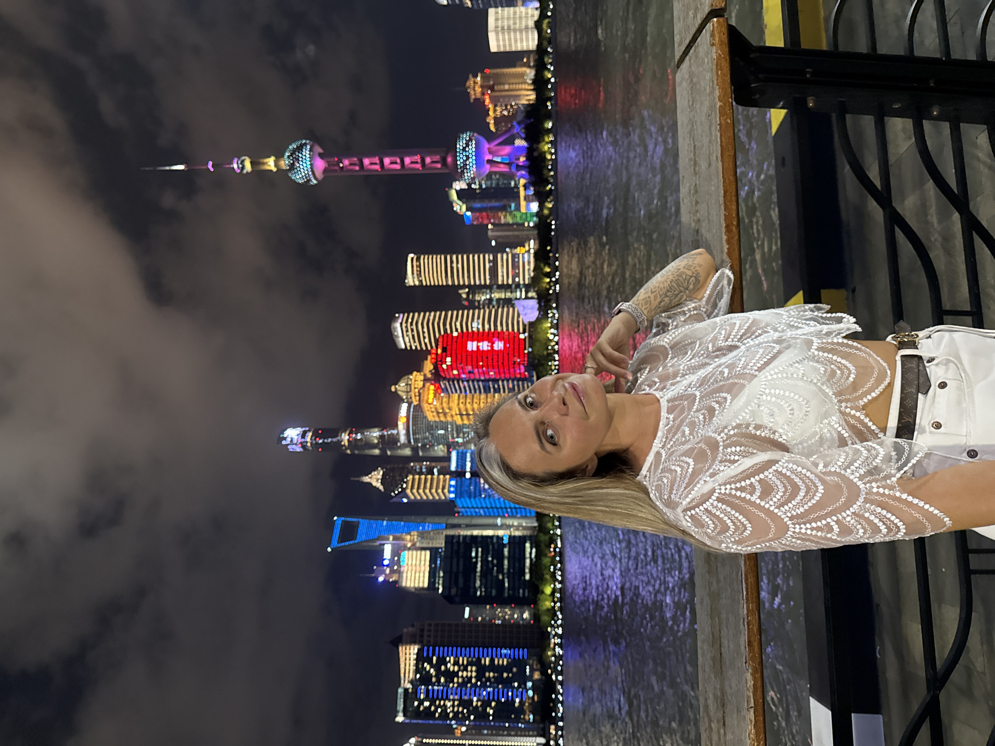 A medium shot captures a woman with blonde hair and light skin standing on a wooden pier at night. She is wearing a white lace crop top and white pants, with a patterned belt. Her left arm is resting on the pier, and her right hand is near her face. Behind her, a body of water reflects the lights of a cityscape. Numerous skyscrapers, illuminated with various colors, dominate the background, including the Oriental Pearl TV Tower. The sky above is dark and cloudy. The scene appears to be a waterfront promenade in Shanghai, likely within the Hongkou District, given the iconic skyline visible across the Huangpu River.