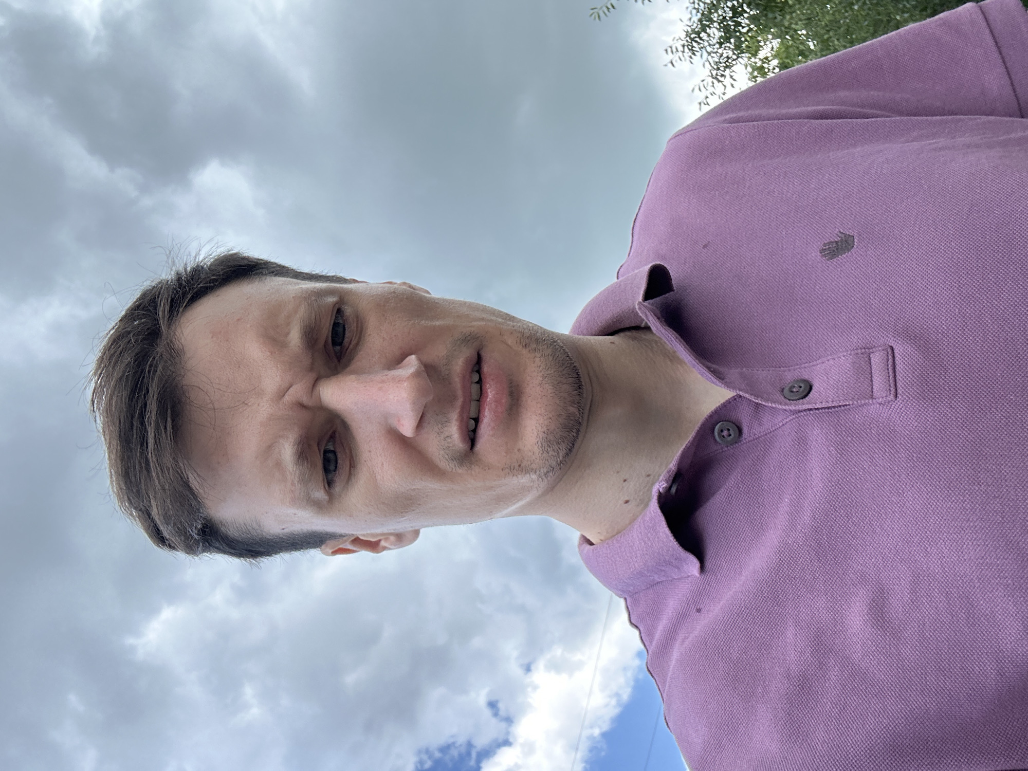 The image shows a close-up, low-angle selfie of a man against a cloudy sky. He is wearing a purple polo shirt with two buttons at the collar. His brow is furrowed, and his mouth is slightly open, suggesting a look of concern or distaste. The background consists of a cloudy sky, with a hint of blue sky and some green foliage visible on the right side. The man's face and upper body fill the frame. The location is stated to be Makiv, Ukraine.