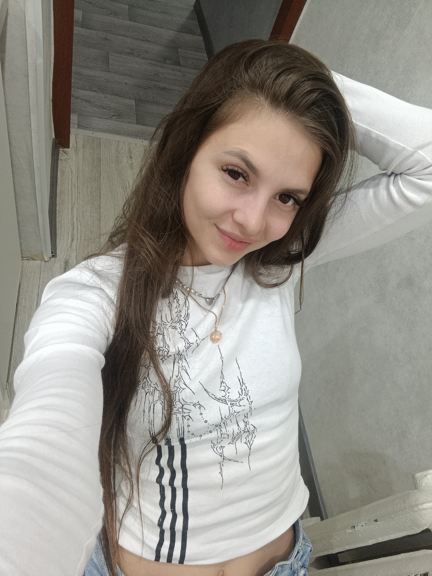 A young woman with long brown hair poses for a selfie. She is wearing a white long-sleeved shirt with a black graphic design and blue denim shorts. She has a necklace and is touching her hair with her left hand, which is visible in the lower left corner. The background shows a wall with wood-paneled and gray flooring, as well as part of a white object on the right side, possibly a railing. The city and specific location are unknown.