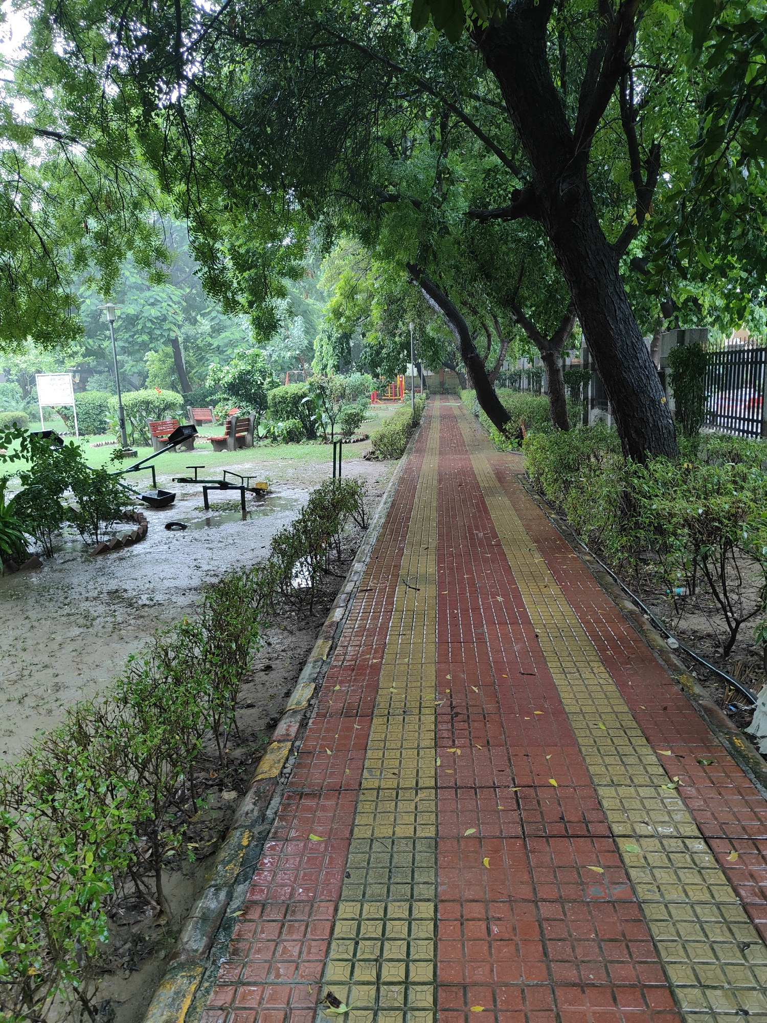 The image depicts a park in Gurgaon, India, after a rainfall. A wet, red and yellow tiled pathway runs diagonally across the frame, bordered by manicured green bushes. To the left of the pathway, a muddy, waterlogged area contains playground equipment, including a see-saw and a swing set, all appearing to be in disuse. Several red benches are visible in the background, along with trees and streetlights. A black metal fence runs along the right side of the pathway, separating the park from the exterior. The overall impression is one of a tranquil, rain-dampened public space.