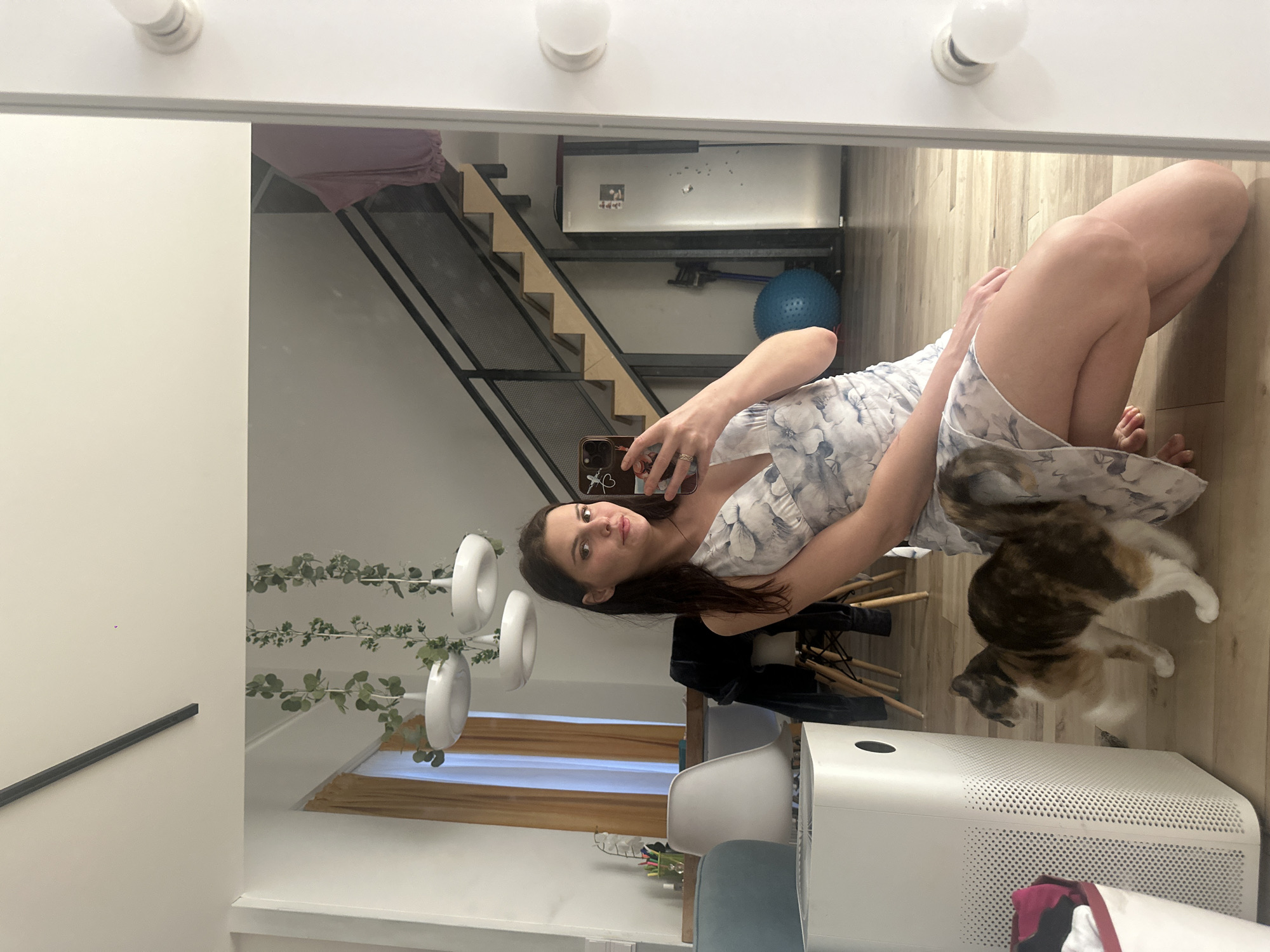 A young woman is taking a selfie in a mirror. She is wearing a light-colored dress with a floral pattern. She is sitting on the floor with her knees bent and her legs crossed. A calico cat is sitting next to her, also looking towards the camera. The room appears to be a living space with a wooden floor and a large mirror that reflects the scene. In the background, there is a staircase with a metal railing and wooden steps. There is also a blue exercise ball and some shelving units with various items. Greenery is hanging from the ceiling. The lighting suggests it is daytime. The woman has a slight smile on her face. There is no visible text in the image.